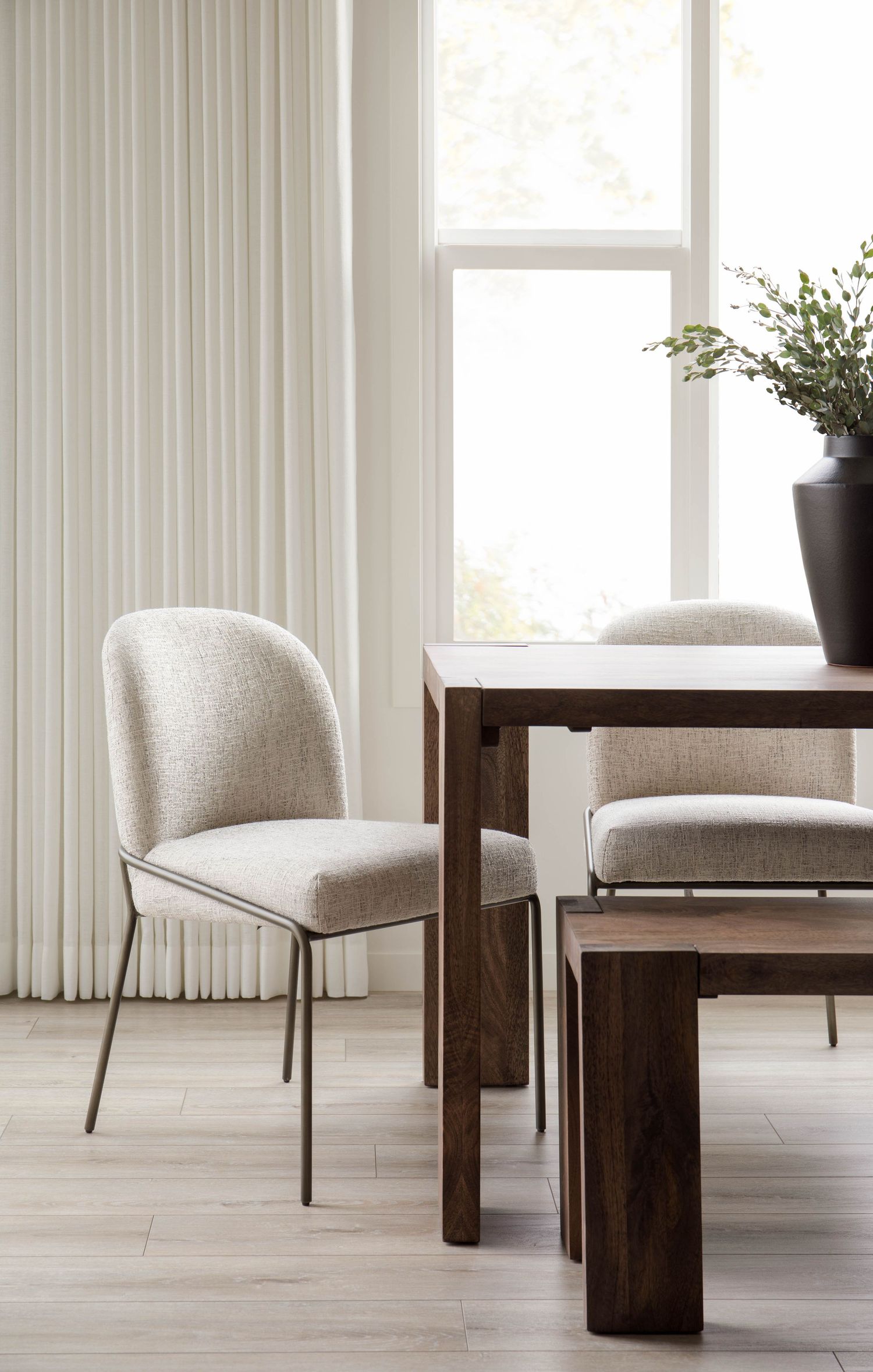 Minimalist dining room with cream upholstered chairs, dark wood table, pleated curtains, and potted greenery.