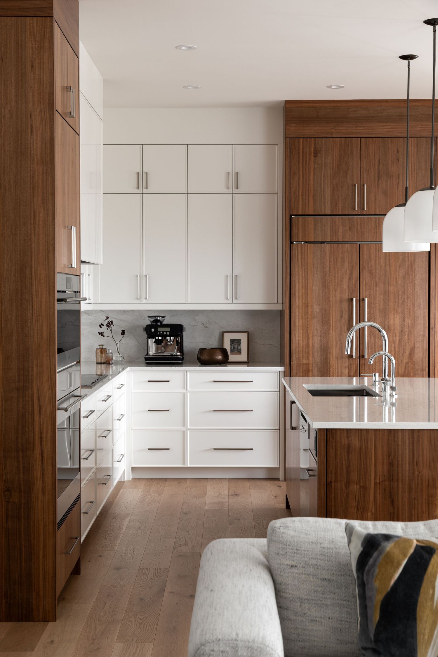 Modern kitchen with white cabinetry, walnut wood accents, gray backsplash, and light hardwood floors in an open-plan layout.