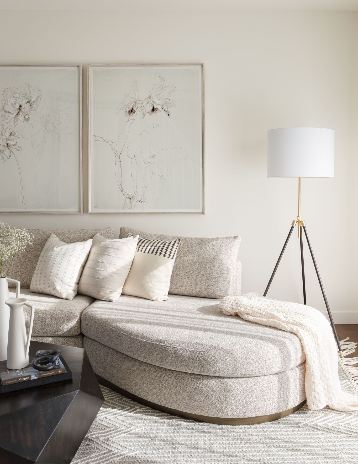 Serene living room with cream curved sofa, oval ottoman, botanical art prints, and tripod floor lamp.