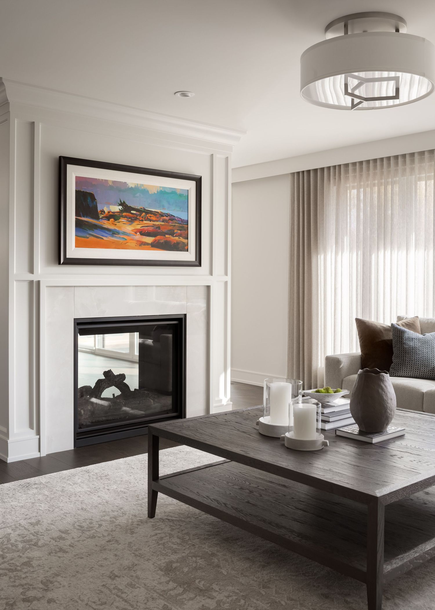 Elegant living room with white fireplace, colorful landscape art, drum ceiling light, and dark wood coffee table.