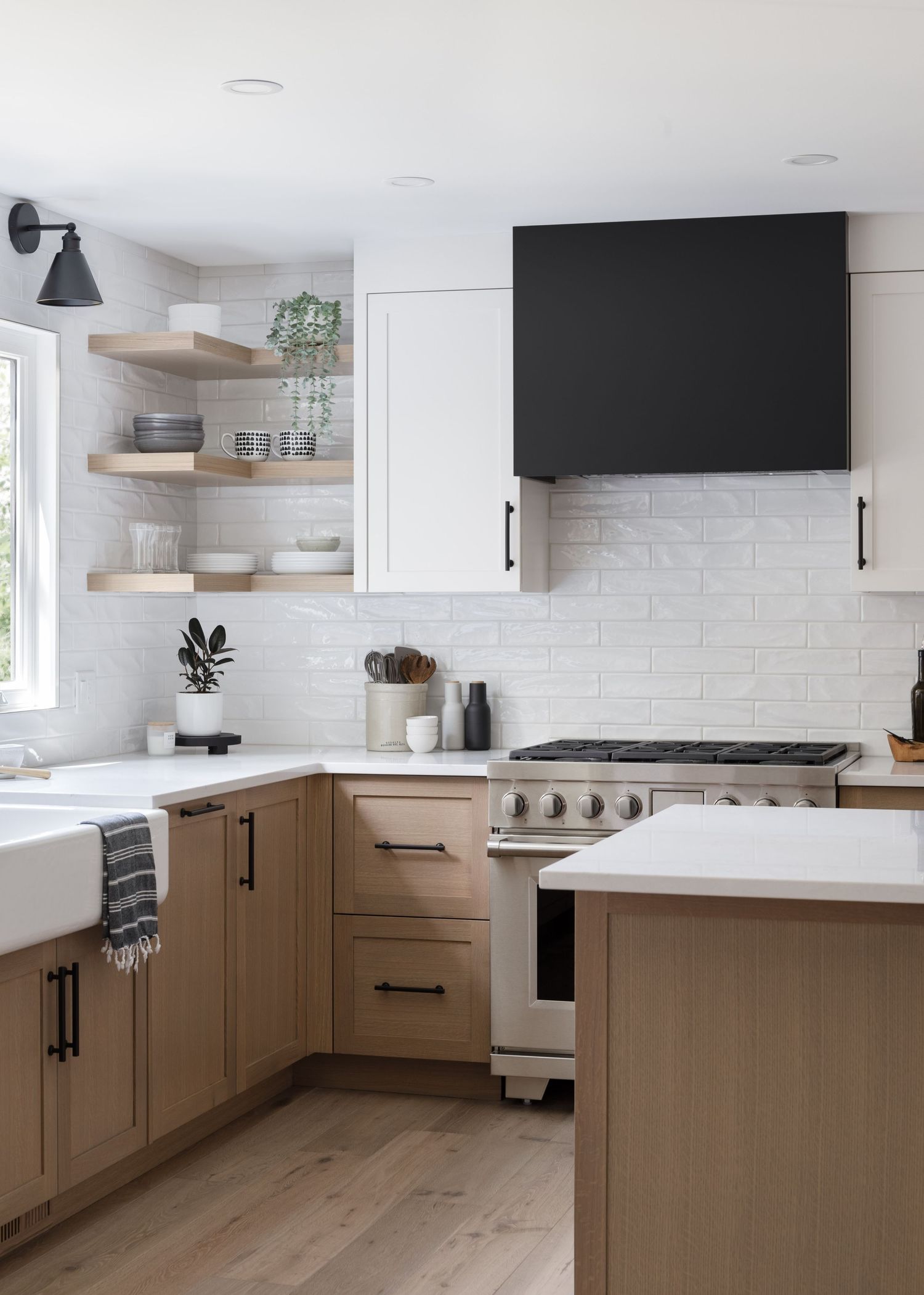 Modern kitchen with light wood lower cabinets, white uppers, open shelves, black range hood, and white subway tile backsplash.
