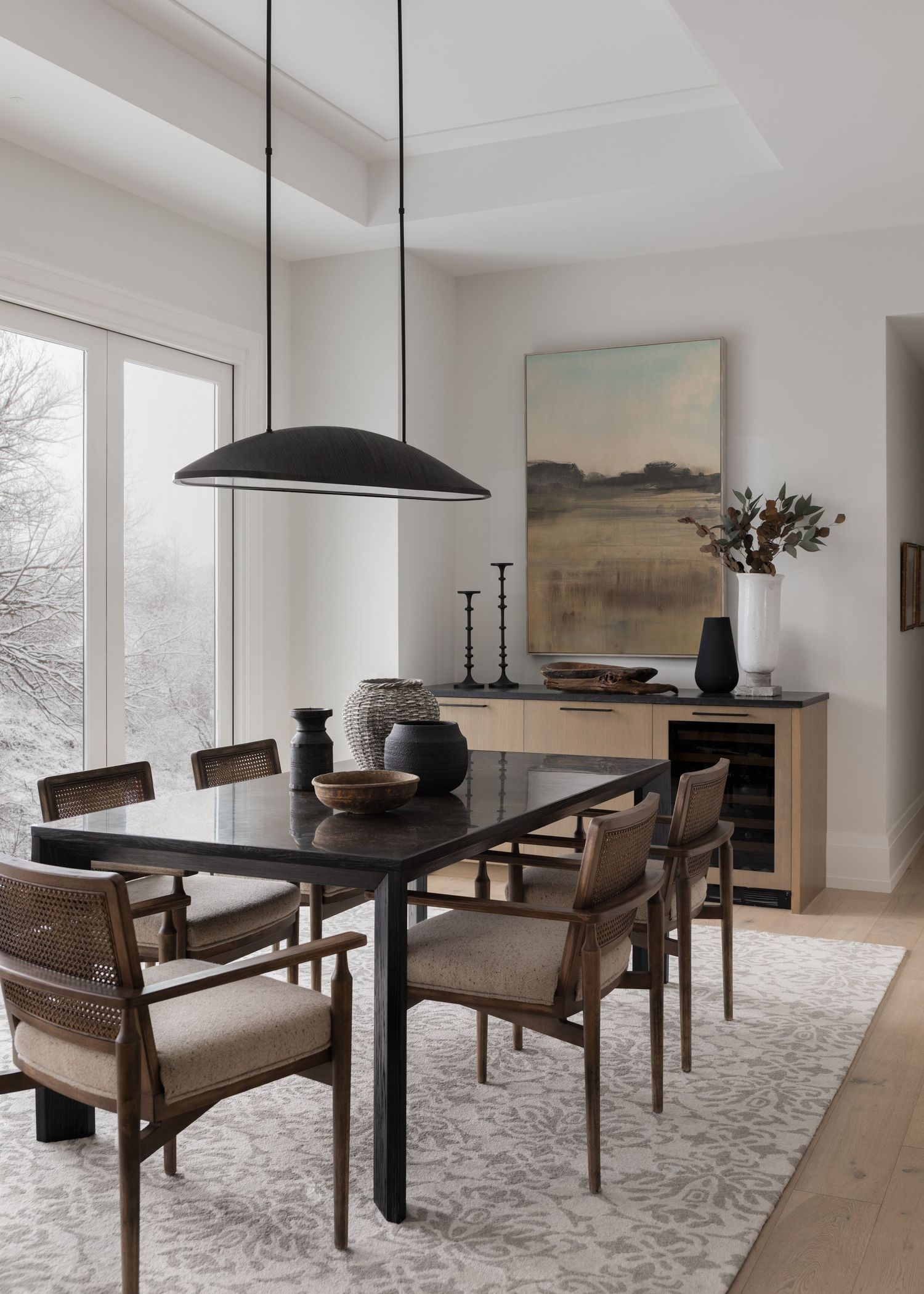Sophisticated dining room with black oval pendant, dark wood table, cane chairs, and landscape artwork above sideboard.