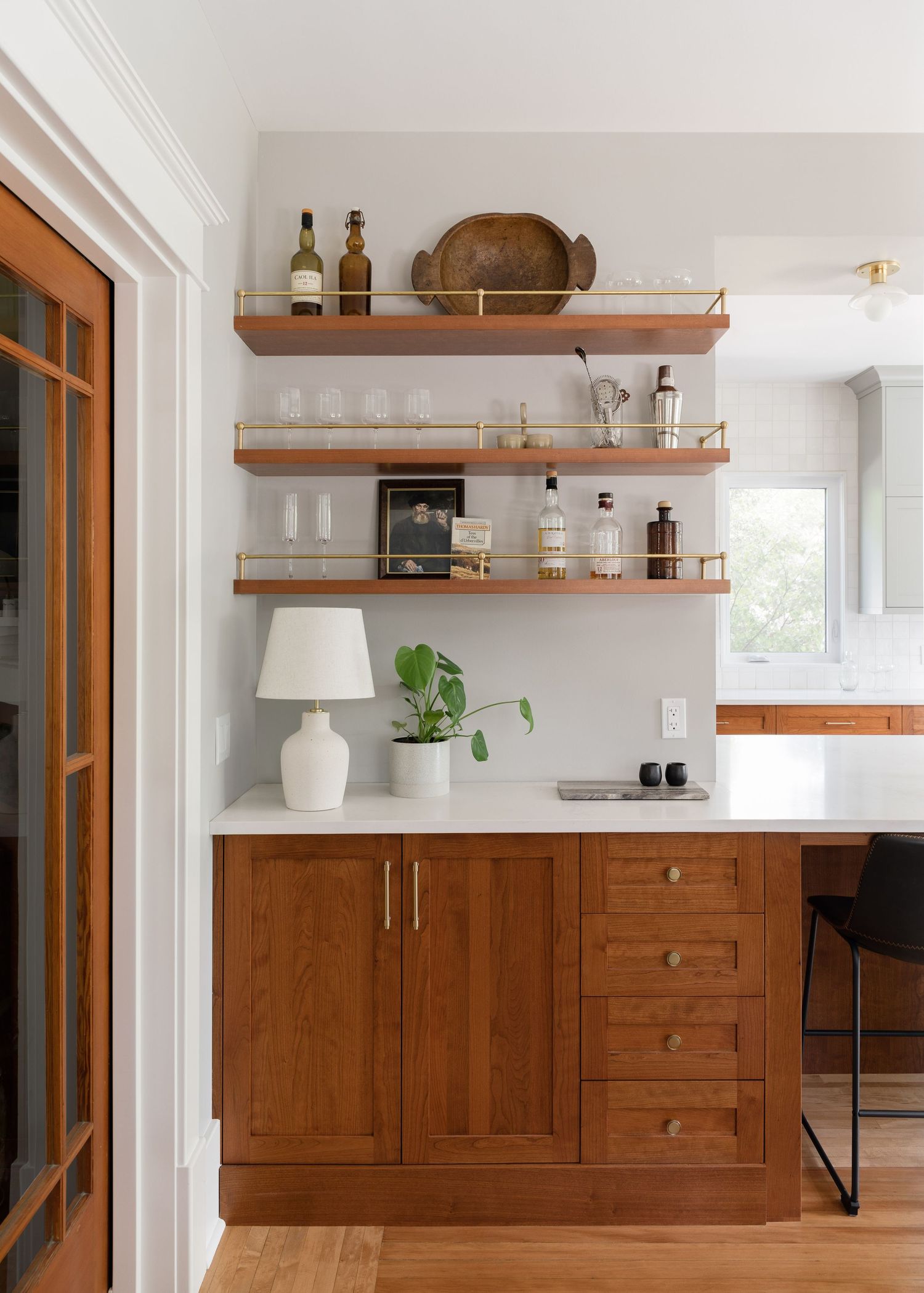 Warm walnut butler pantry with open brass shelving displaying barware and bottles above cabinets with drawers and white countertop.