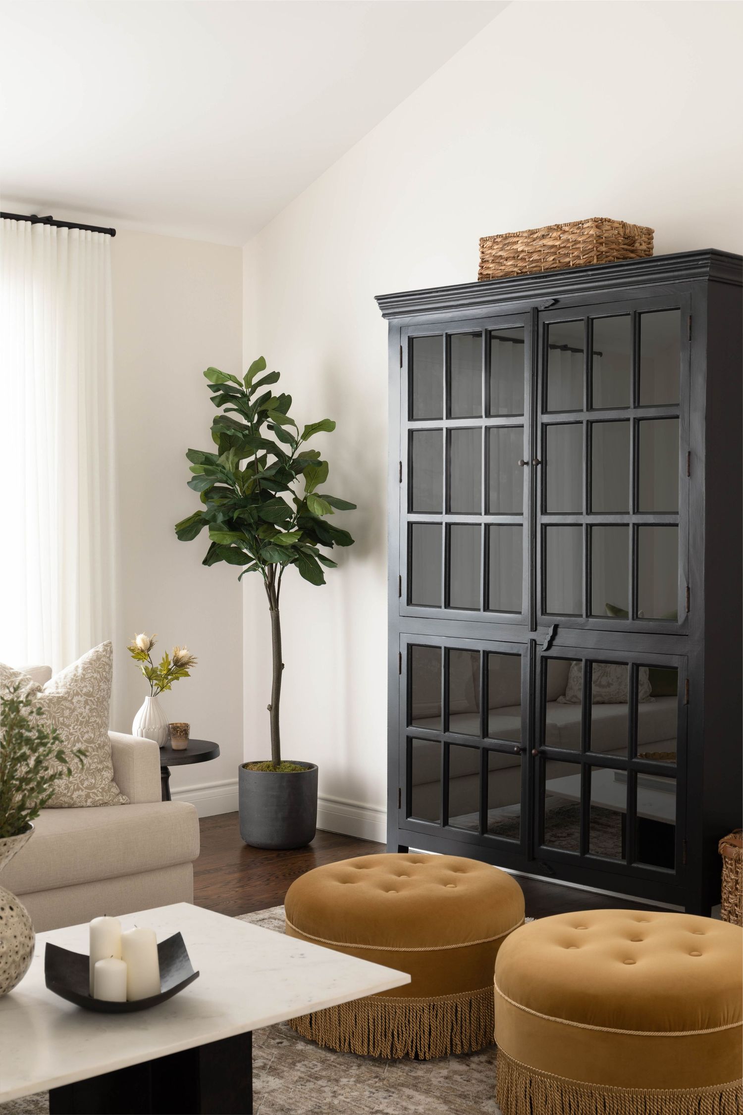 Airy living room with tall black glass cabinet, fiddle leaf fig tree, mustard tufted ottomans, and white sofa.