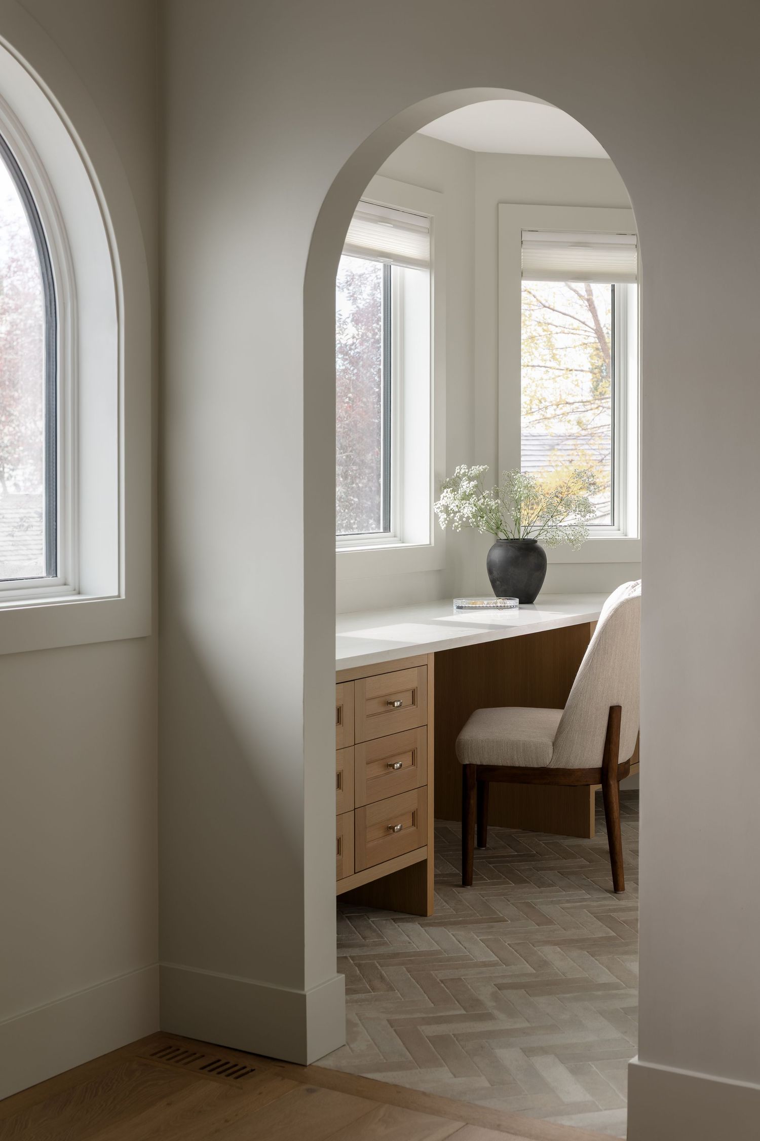 Arched doorway leading to built-in wood desk nook with drawers, neutral chair, and garden windows.