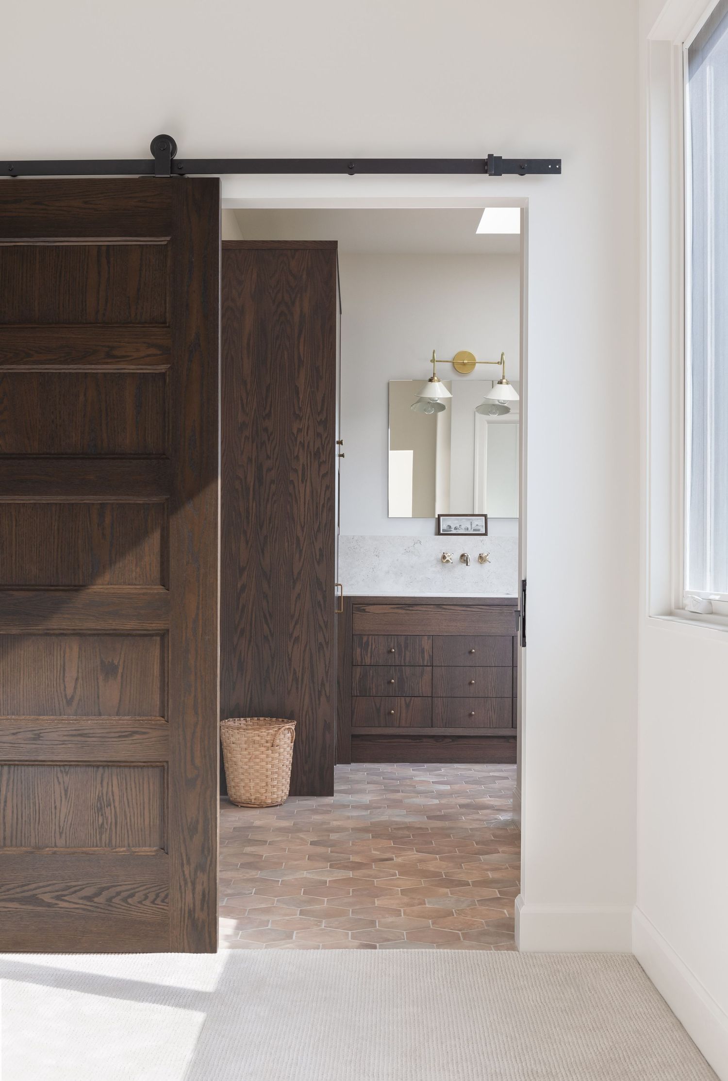 Dark wood sliding barn door opening to a bathroom with marble vanity, brass sconces, and terracotta tile floor.