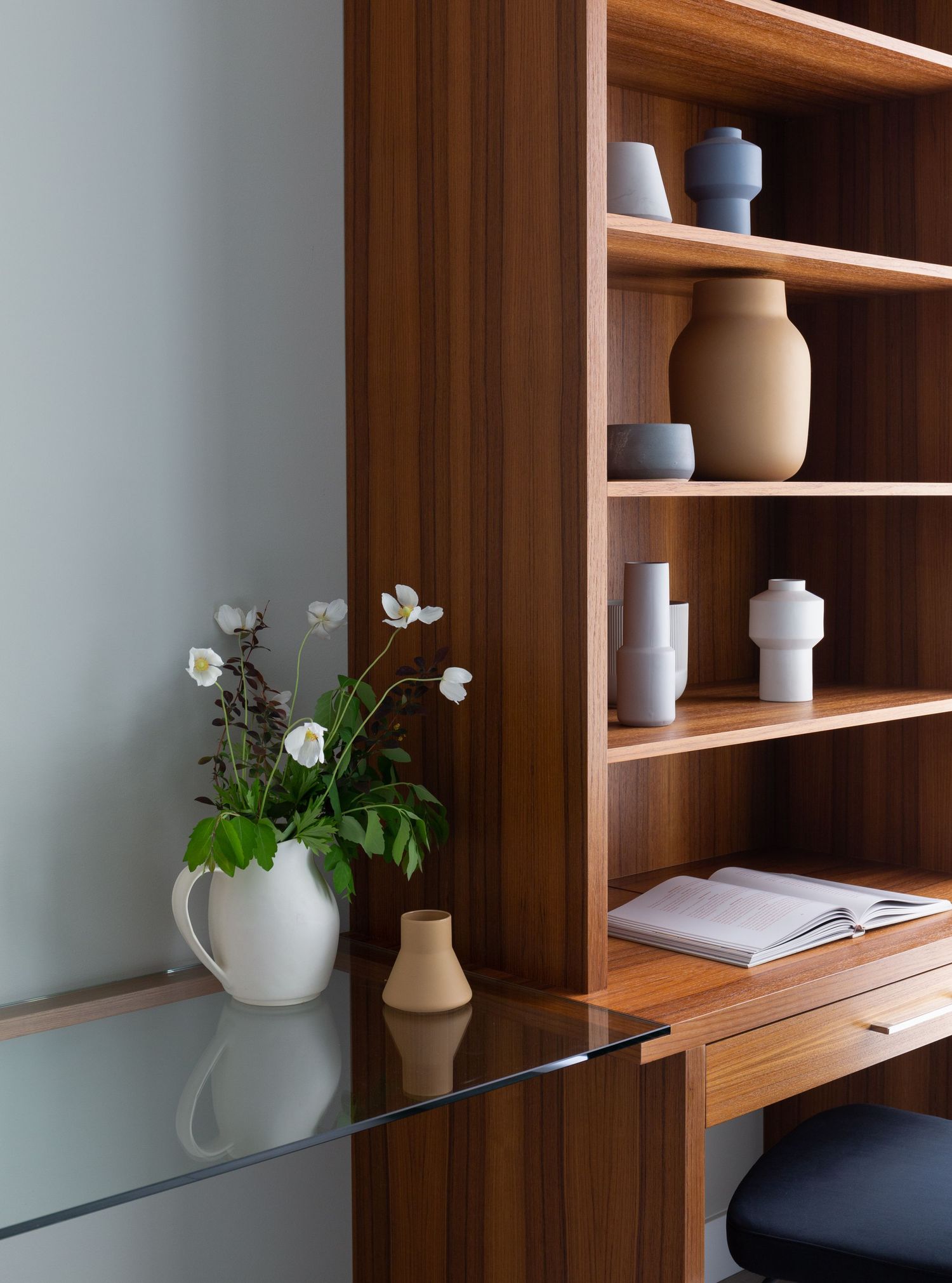 Modern walnut built-in shelving unit displaying ceramic vases with a glass desk and white flower arrangement in foreground.