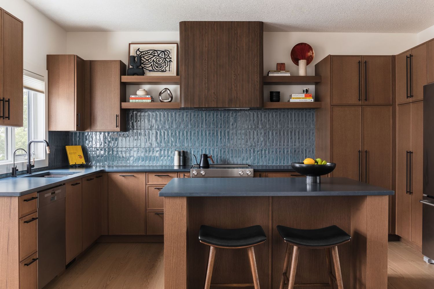 Mid-century modern kitchen featuring walnut cabinetry, blue textured tile backsplash, dark countertops, and island seating.