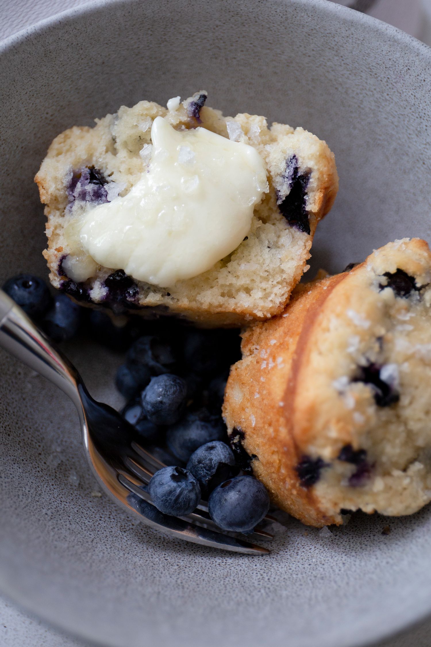 Blueberry muffin split open with butter melting on top, served in a gray bowl with fresh blueberries.
