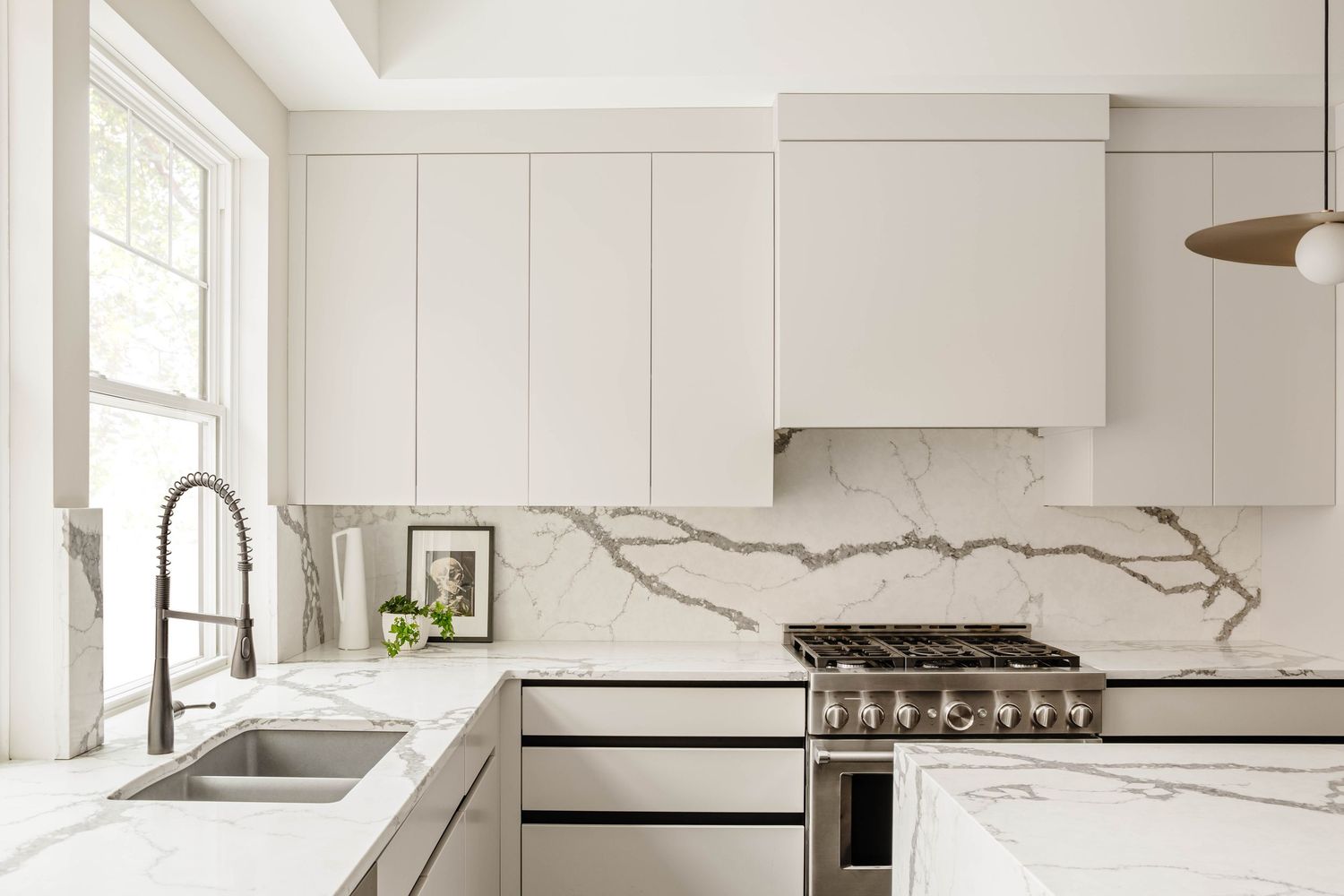 Elegant all-white kitchen featuring marble backsplash, stainless range, farmhouse sink, and flat-front cabinetry.