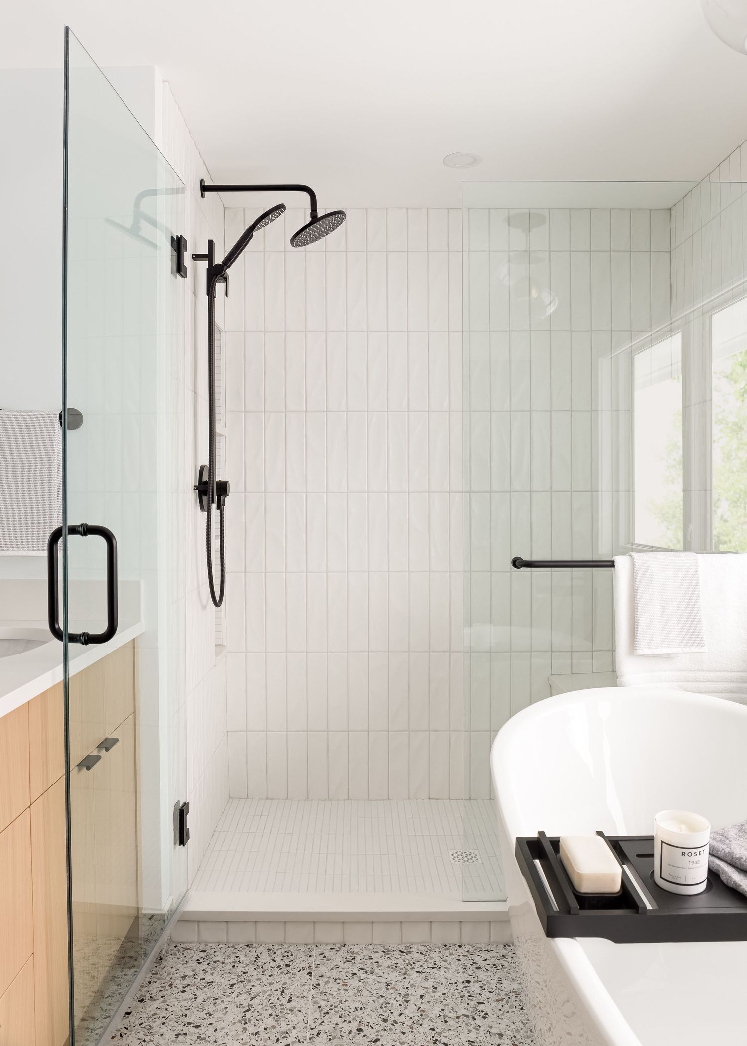 Modern bathroom with black shower fixtures, vertical white tile, terrazzo floor, glass enclosure, and freestanding tub.