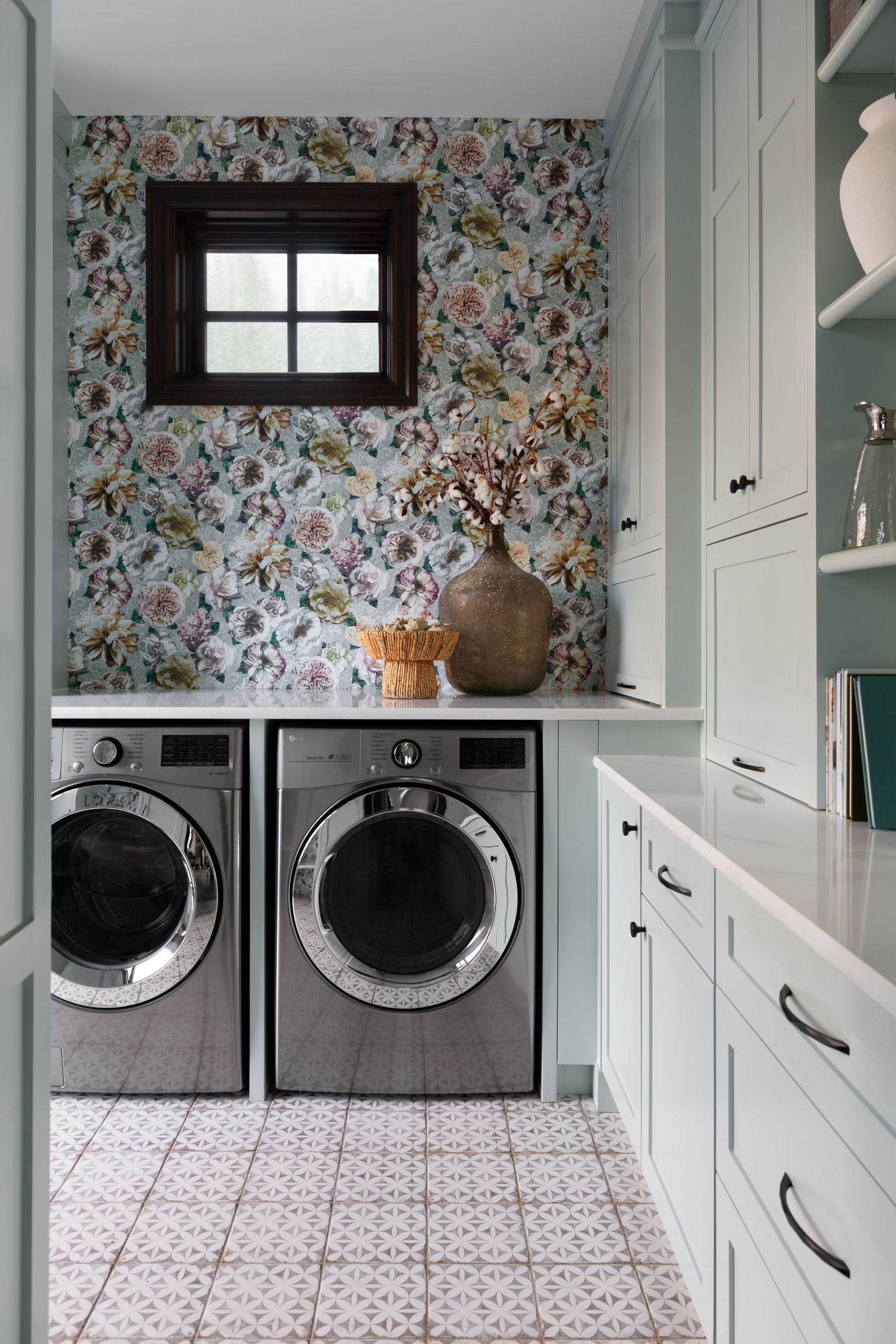 Charming laundry room with floral wallpaper, sage green cabinets, patterned tile floor, and front-load washers.