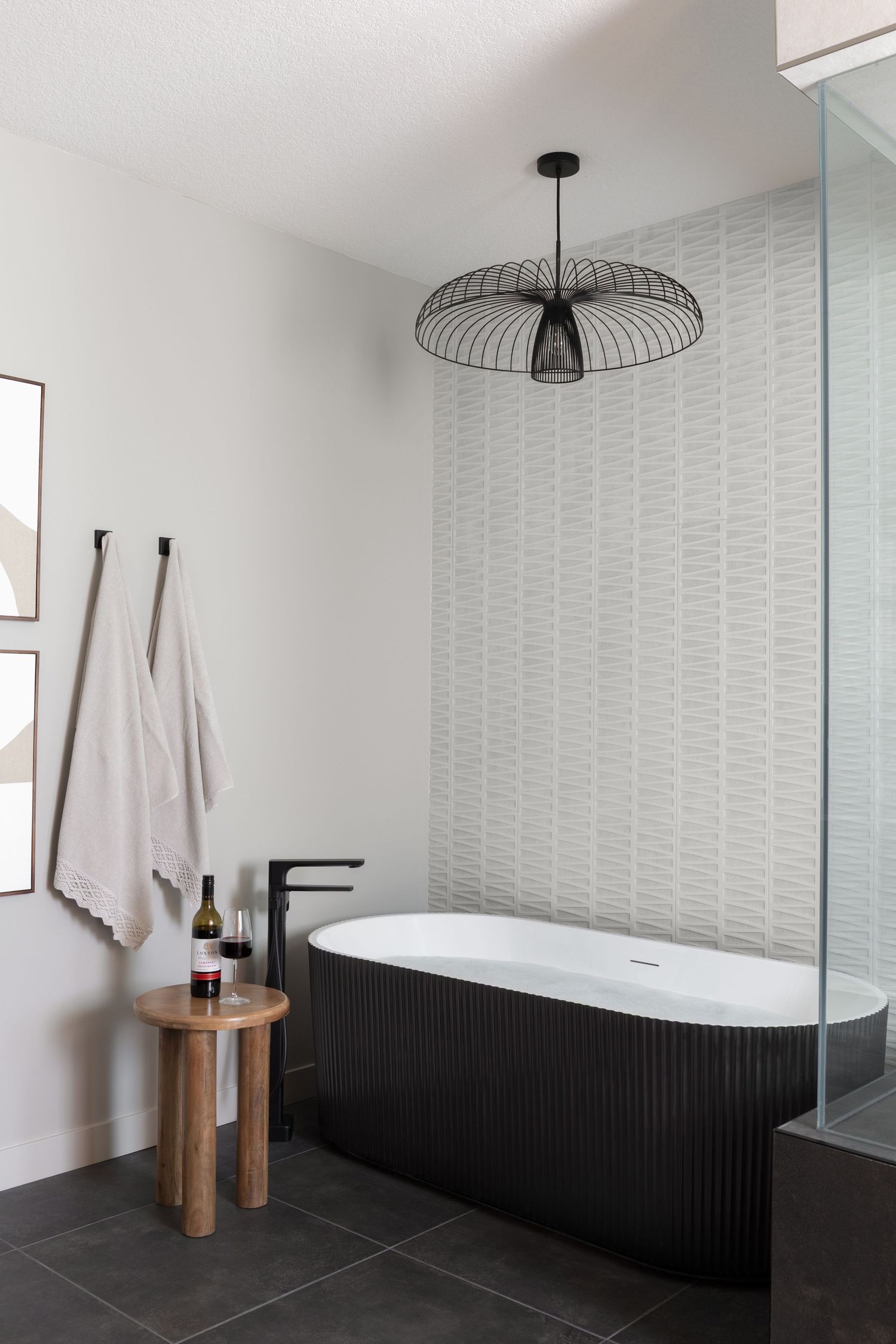 Modern black freestanding bathtub with wire pendant light, wood stool, and textured tile accent wall.