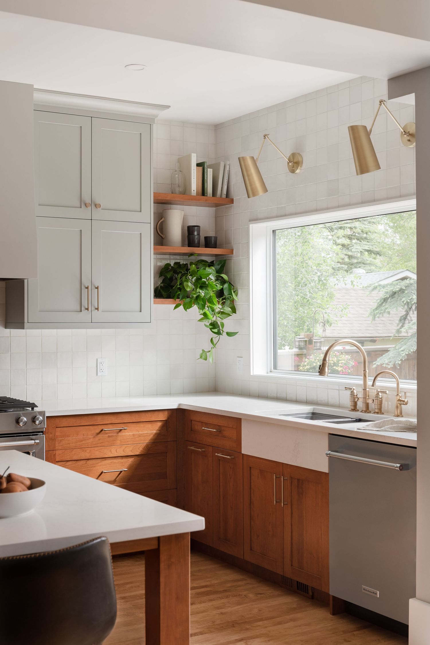 Cozy kitchen with two-tone gray and walnut cabinets, farmhouse sink, brass fixtures, open shelving, and large window with garden view.