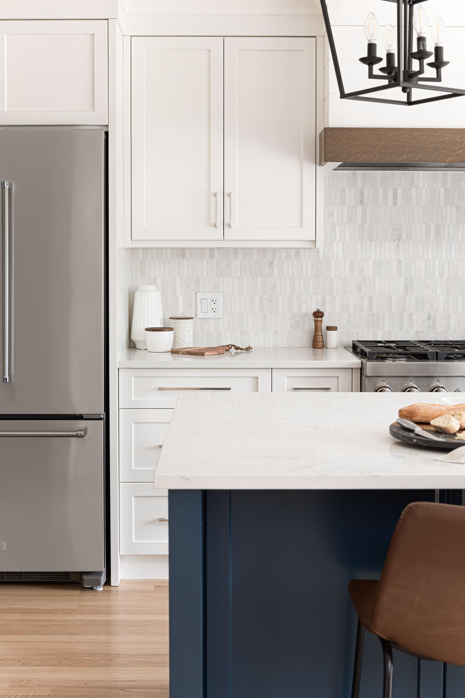 White shaker kitchen with navy island, marble backsplash, stainless appliances, and black chandelier.