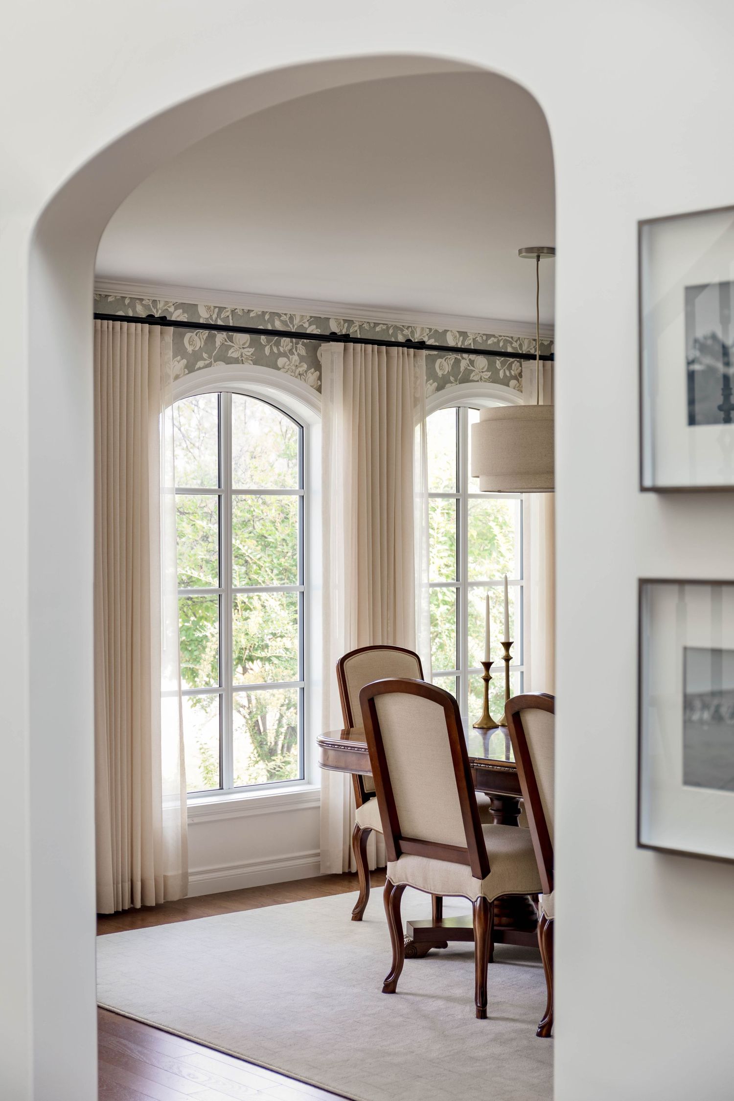 Arched doorway framing dining room with tall arched windows, cream curtains, and upholstered wood chairs.