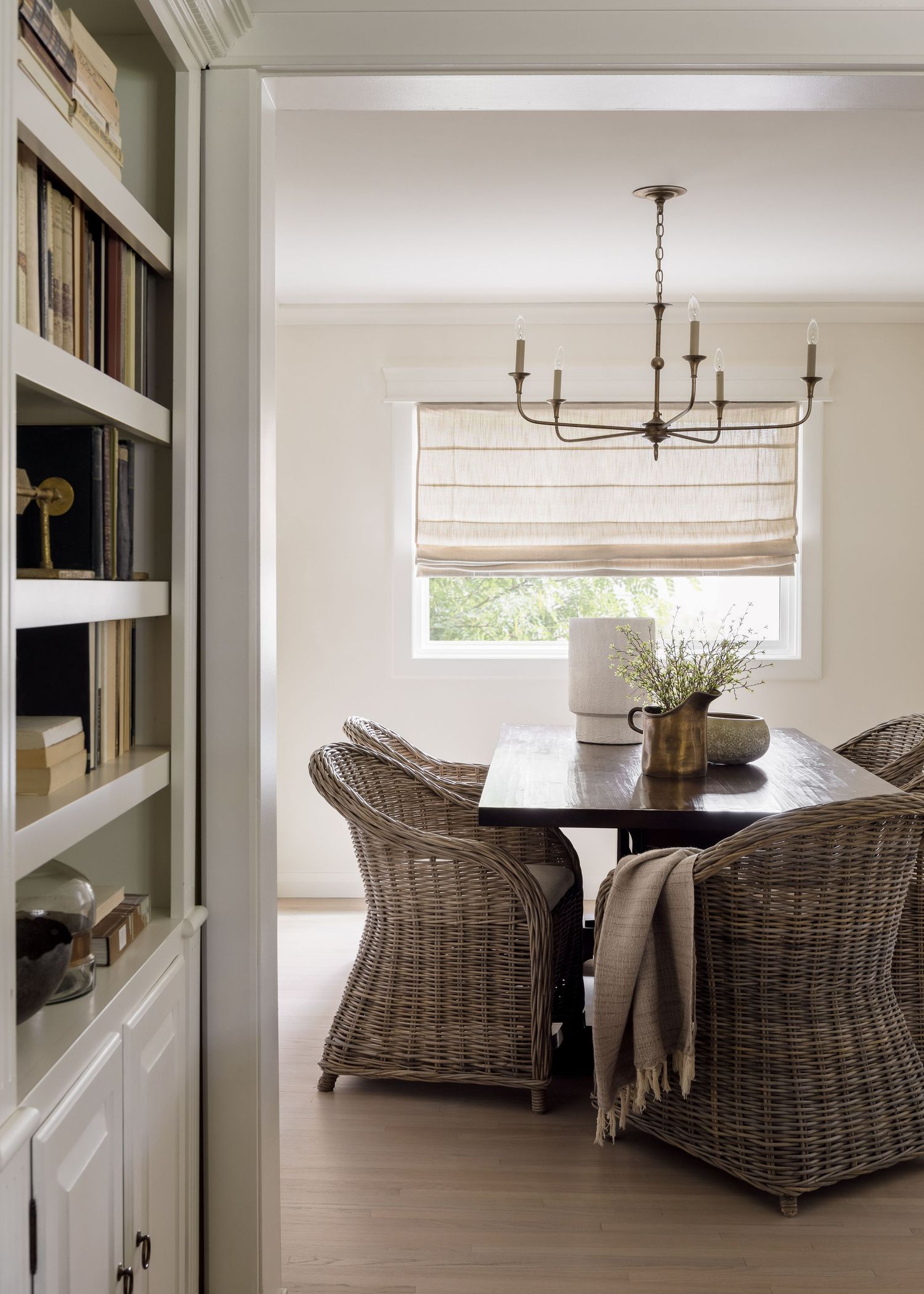 Neutral dining room with wicker chairs, dark wood table, Roman shade, and antique brass chandelier.