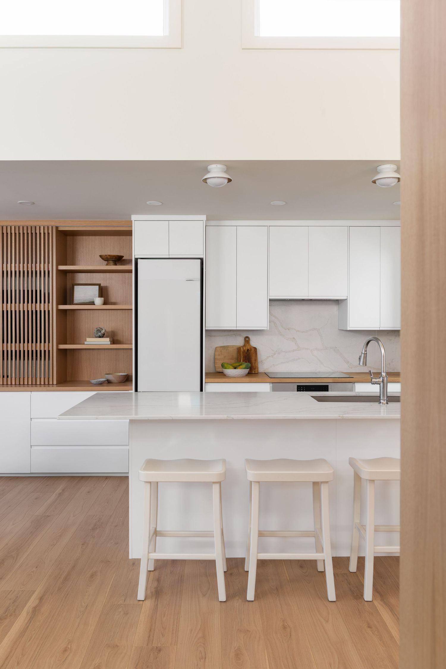 Modern white kitchen with marble island, three wooden stools, white cabinets, and natural wood open shelving unit.