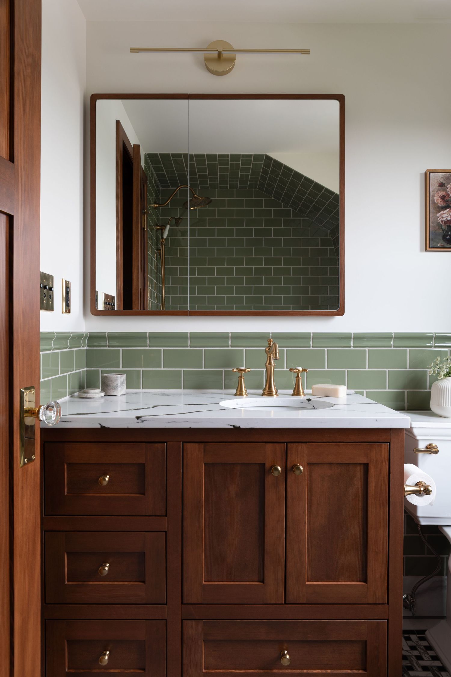 Dark wood bathroom vanity with marble countertop, brass faucet, and green subway tile backsplash with framed mirror.
