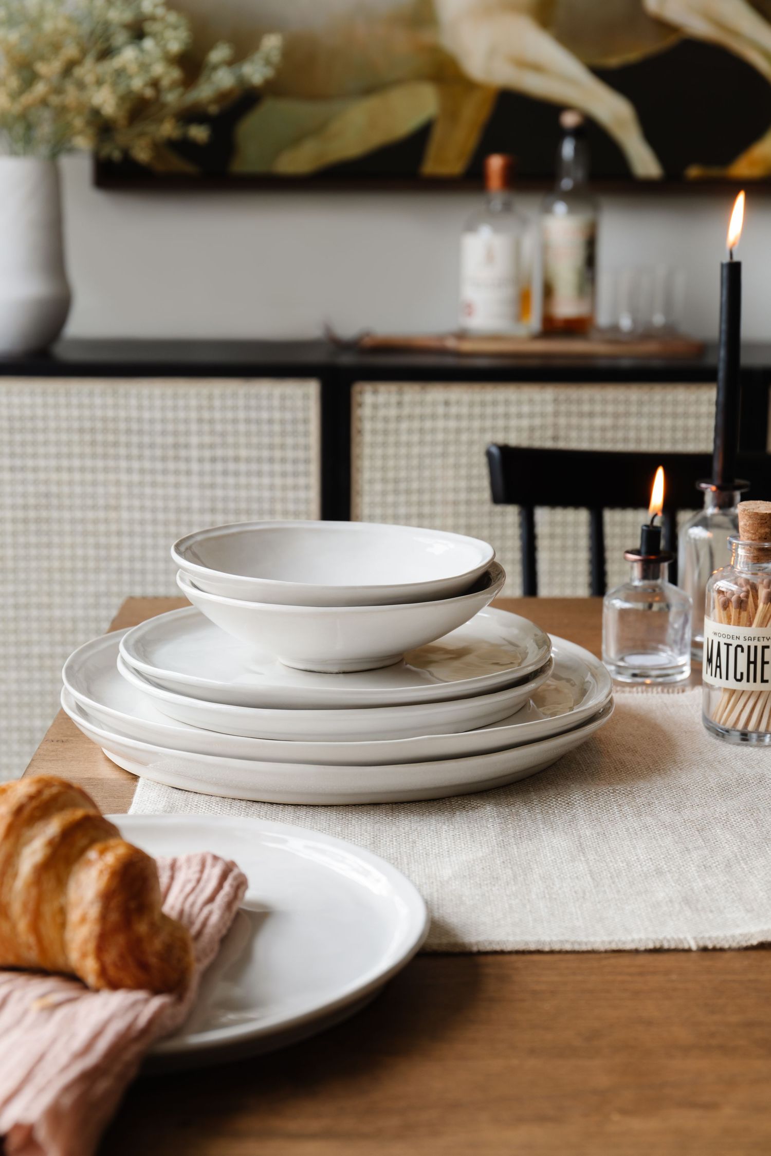 Elegant white ceramic dinnerware set stacked on wooden table with candles and croissant in cozy setting.
