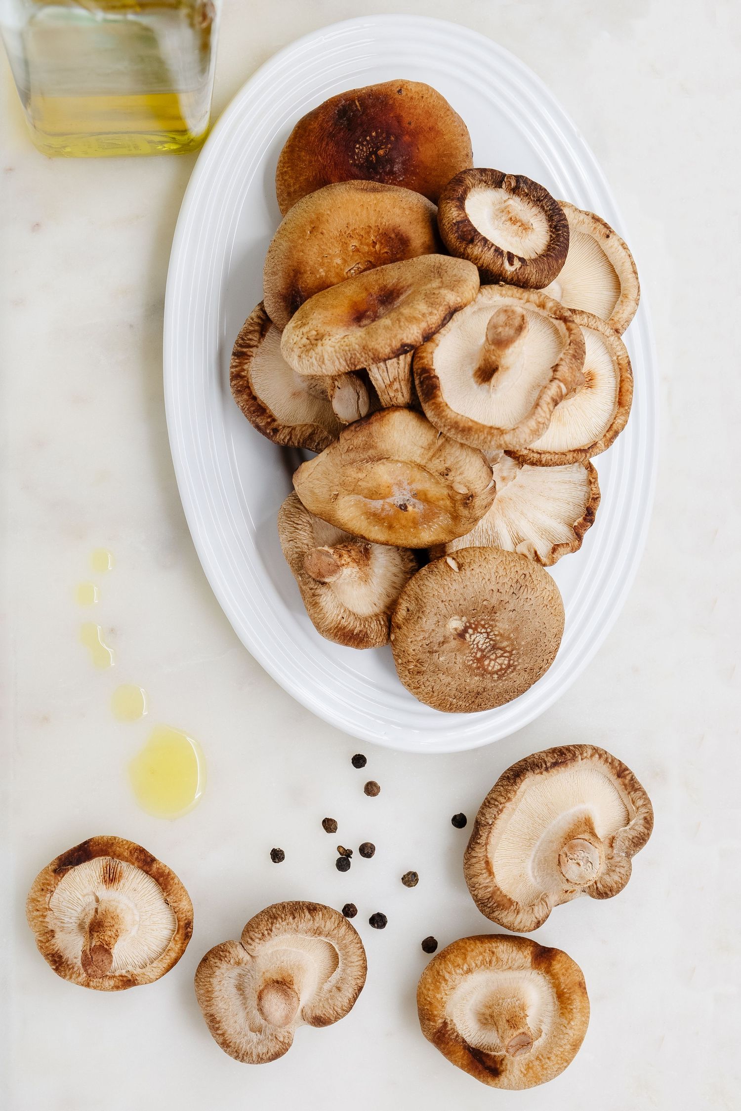 Fresh shiitake mushrooms on a white oval plate with olive oil and black peppercorns on marble surface.