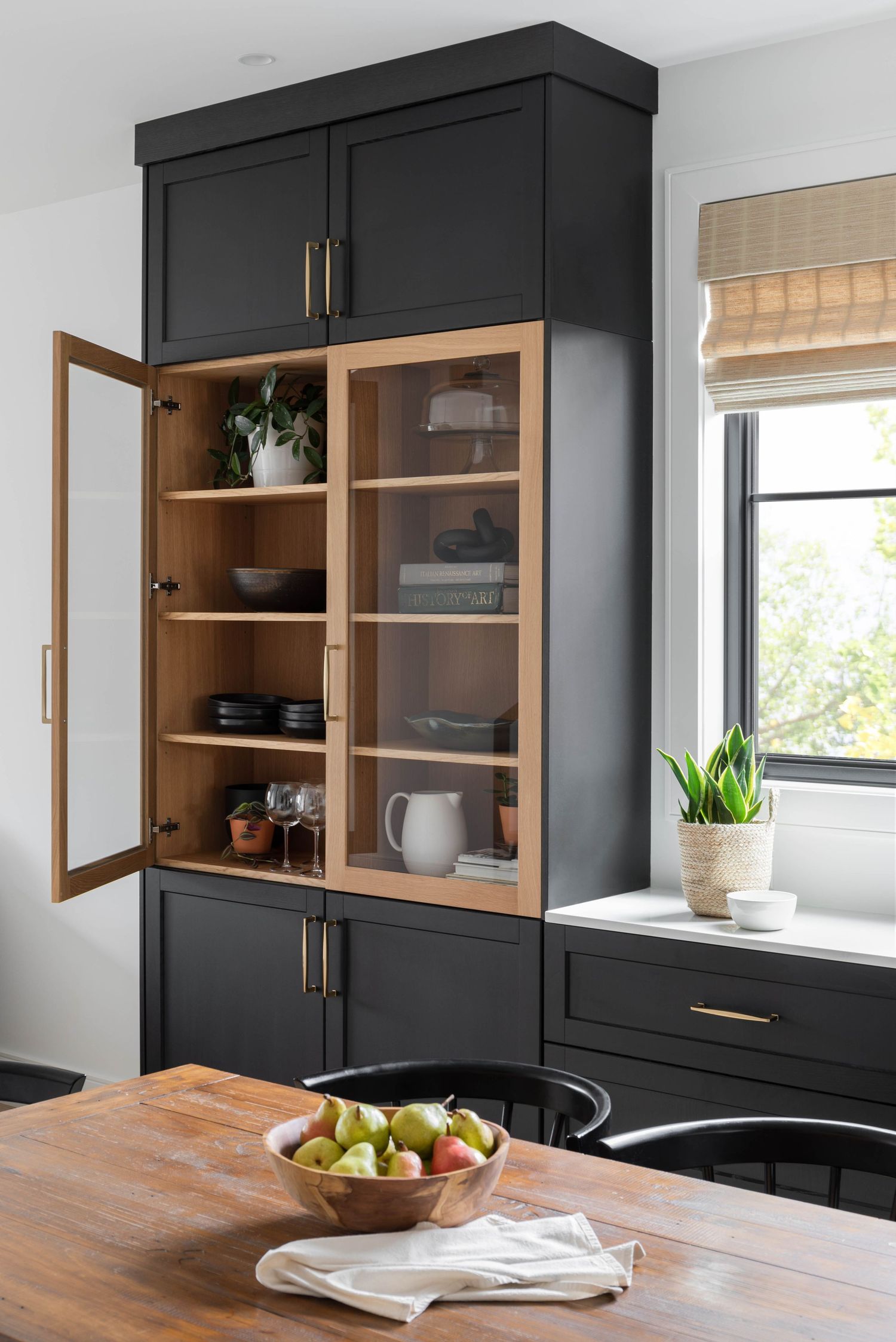 Bold black kitchen cabinet with open wood-interior glass doors displaying dishware, beside a wood dining table with fruit bowl.