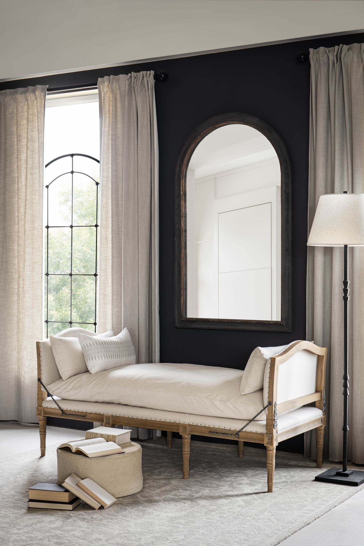 Elegant bedroom sitting area with cream chaise lounge, black accent wall, arched mirror, and tall arched window.