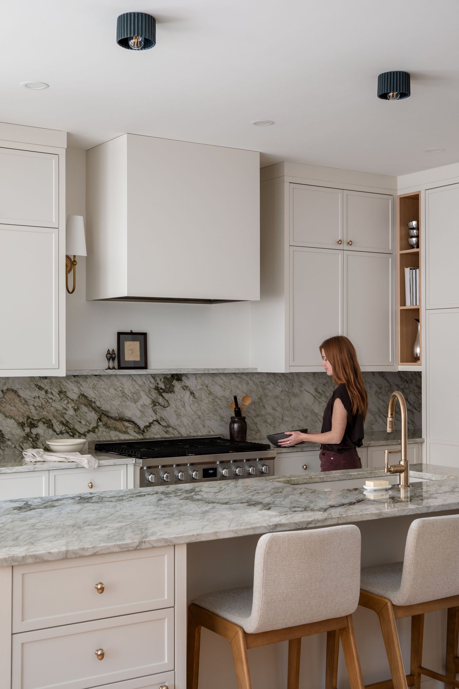 Woman cooking in elegant white kitchen with marble countertops, brass fixtures, and custom range hood.