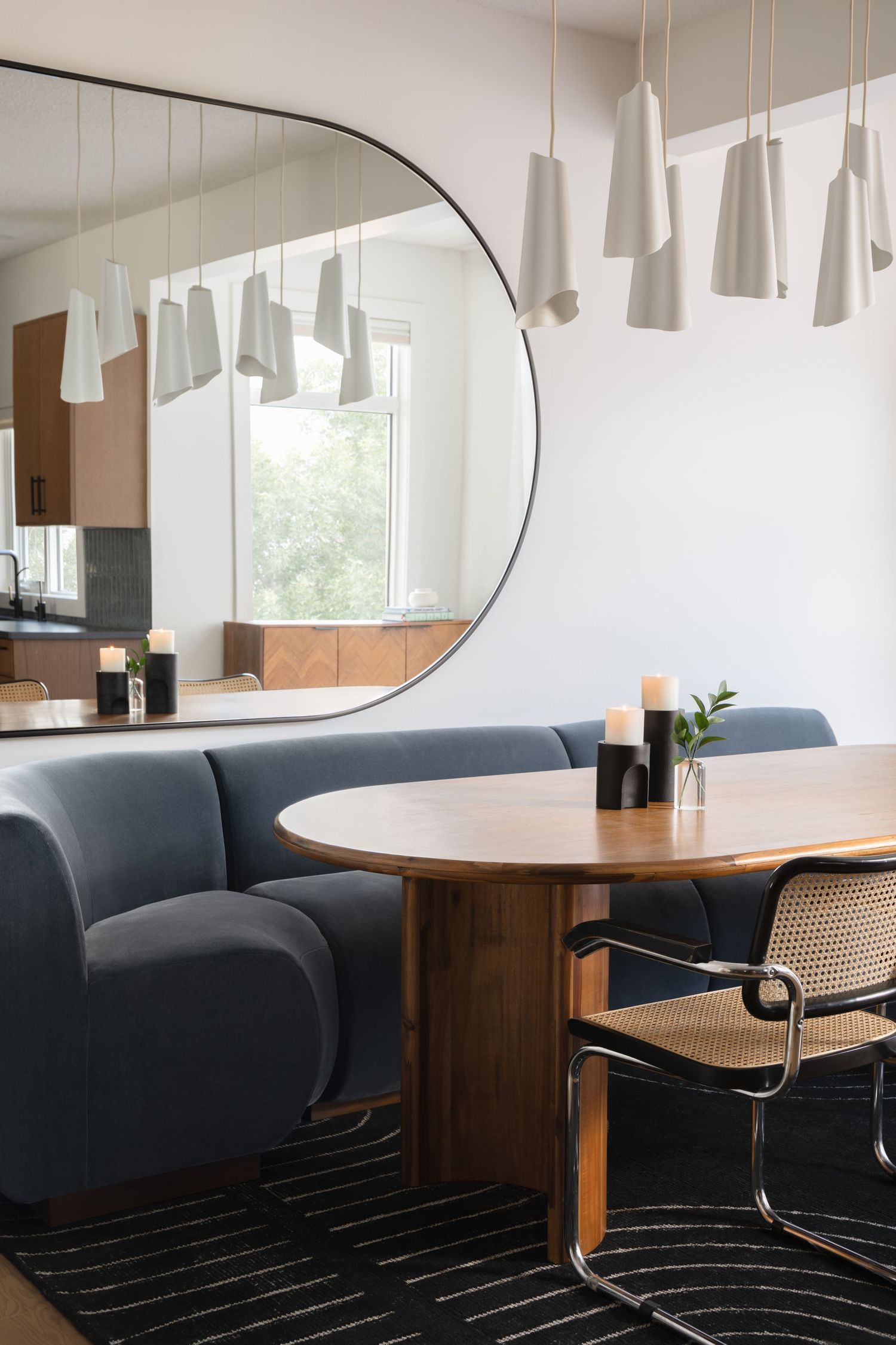 Modern dining nook with curved blue velvet banquette, round wood table, cane chairs, and large arch mirror.