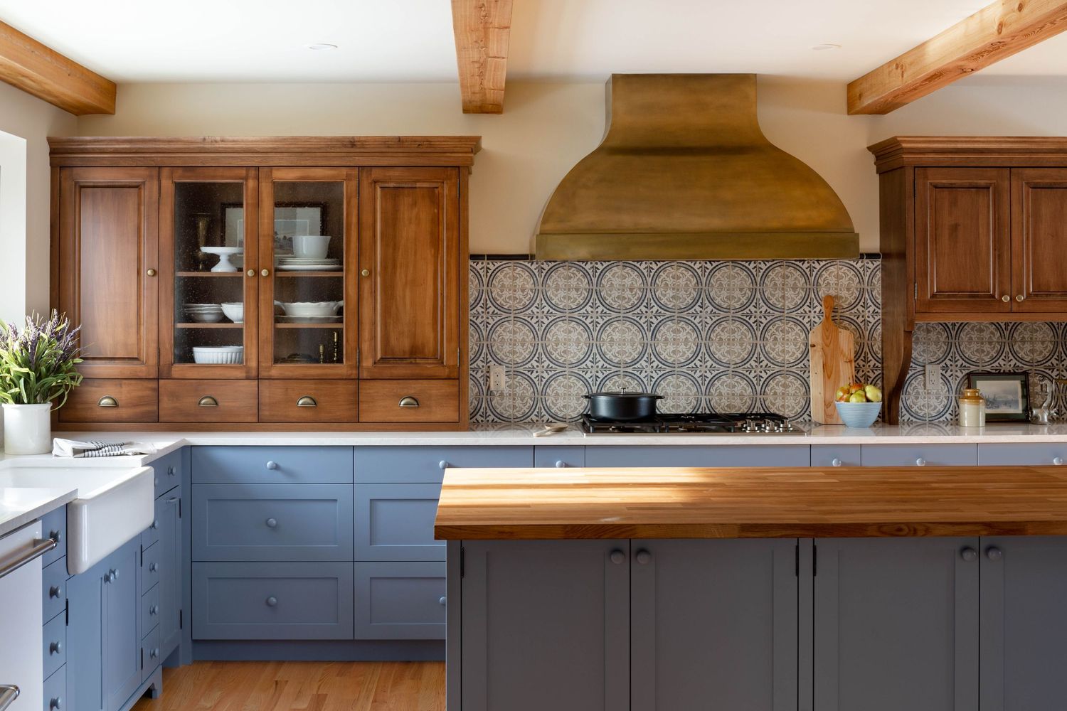 Rustic kitchen with blue shaker cabinets, wood island, ornate tile backsplash, brass range hood, and exposed wooden ceiling beams.