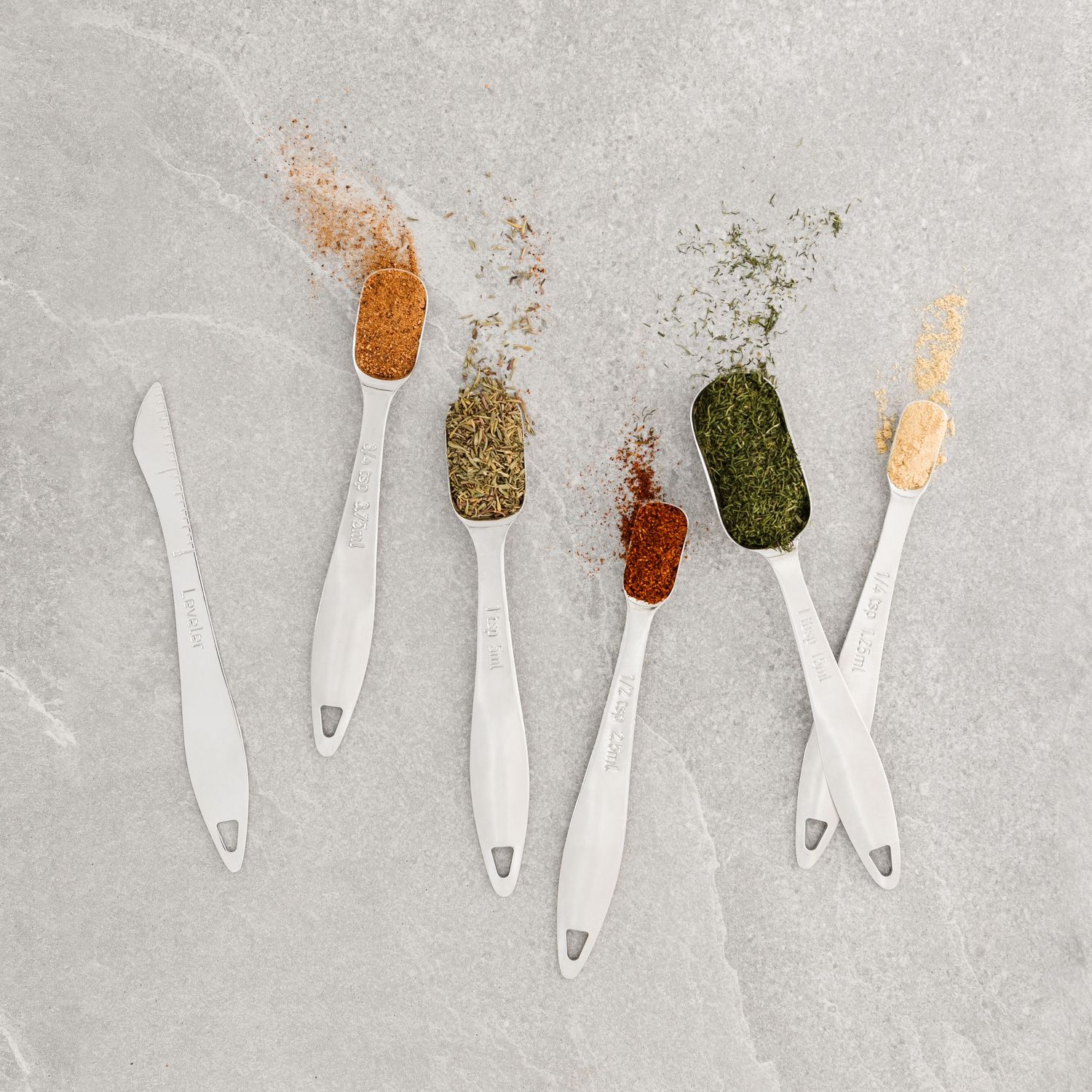Five white measuring spoons with various colorful spices including paprika, herbs, and seasoning blends on marble surface.