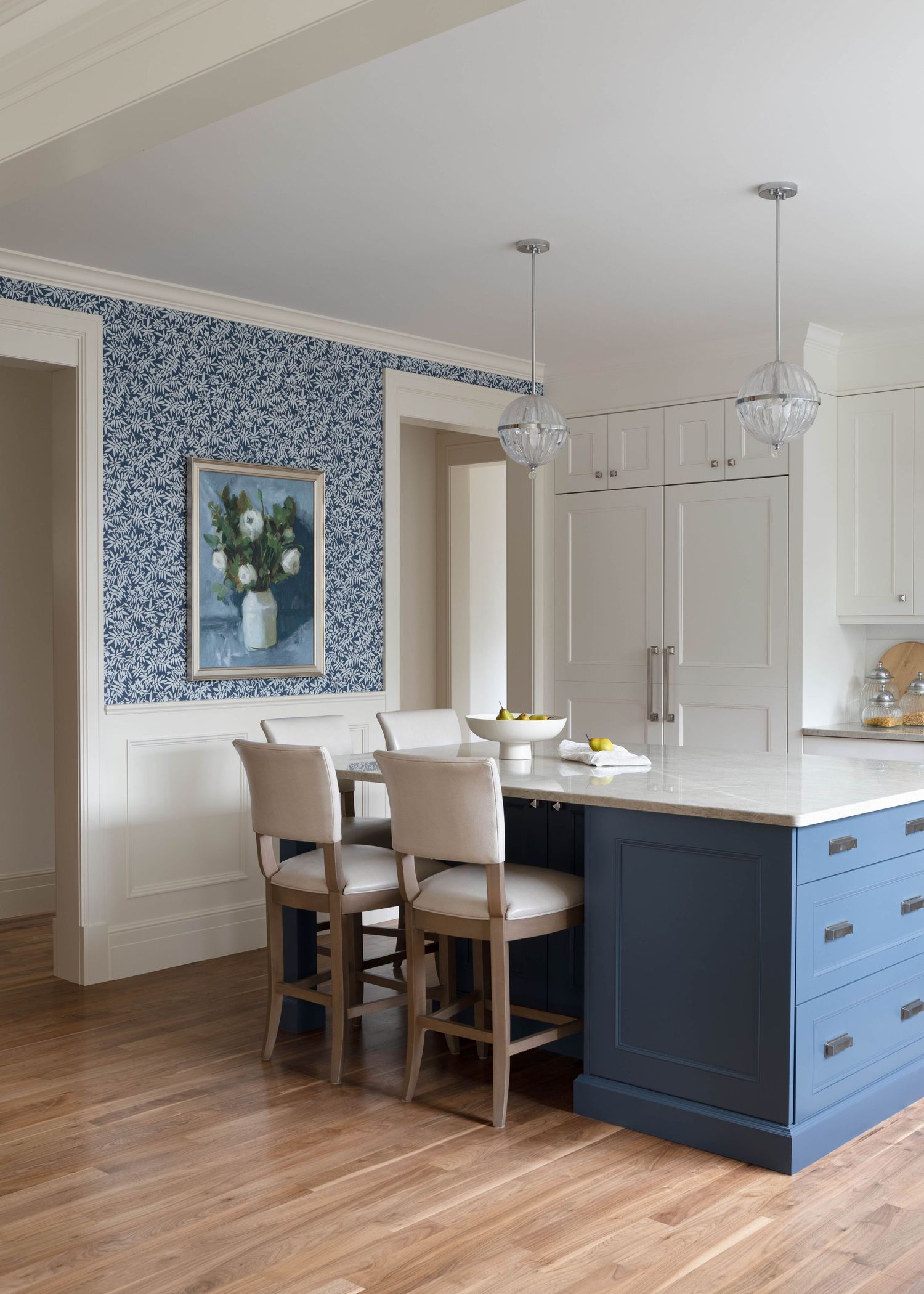 Blue and white kitchen with navy island, pendant lights, upholstered barstools, and floral wallpaper accent wall.
