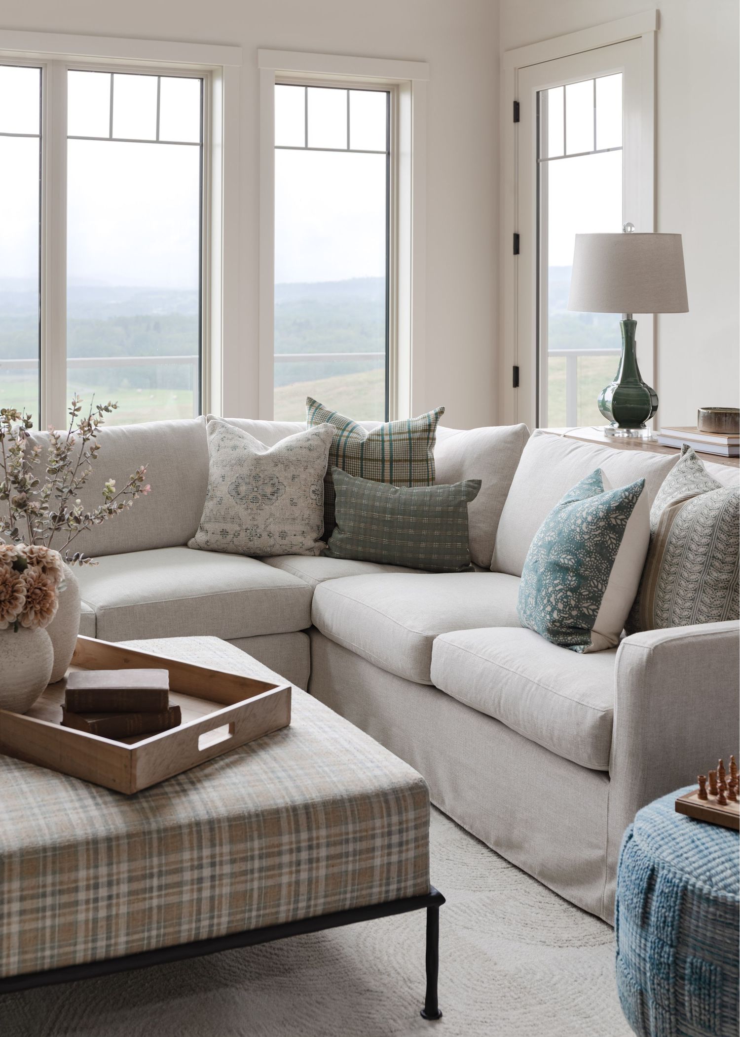 Cozy farmhouse living room with linen sectional, plaid ottoman, layered throw pillows, and scenic window views.