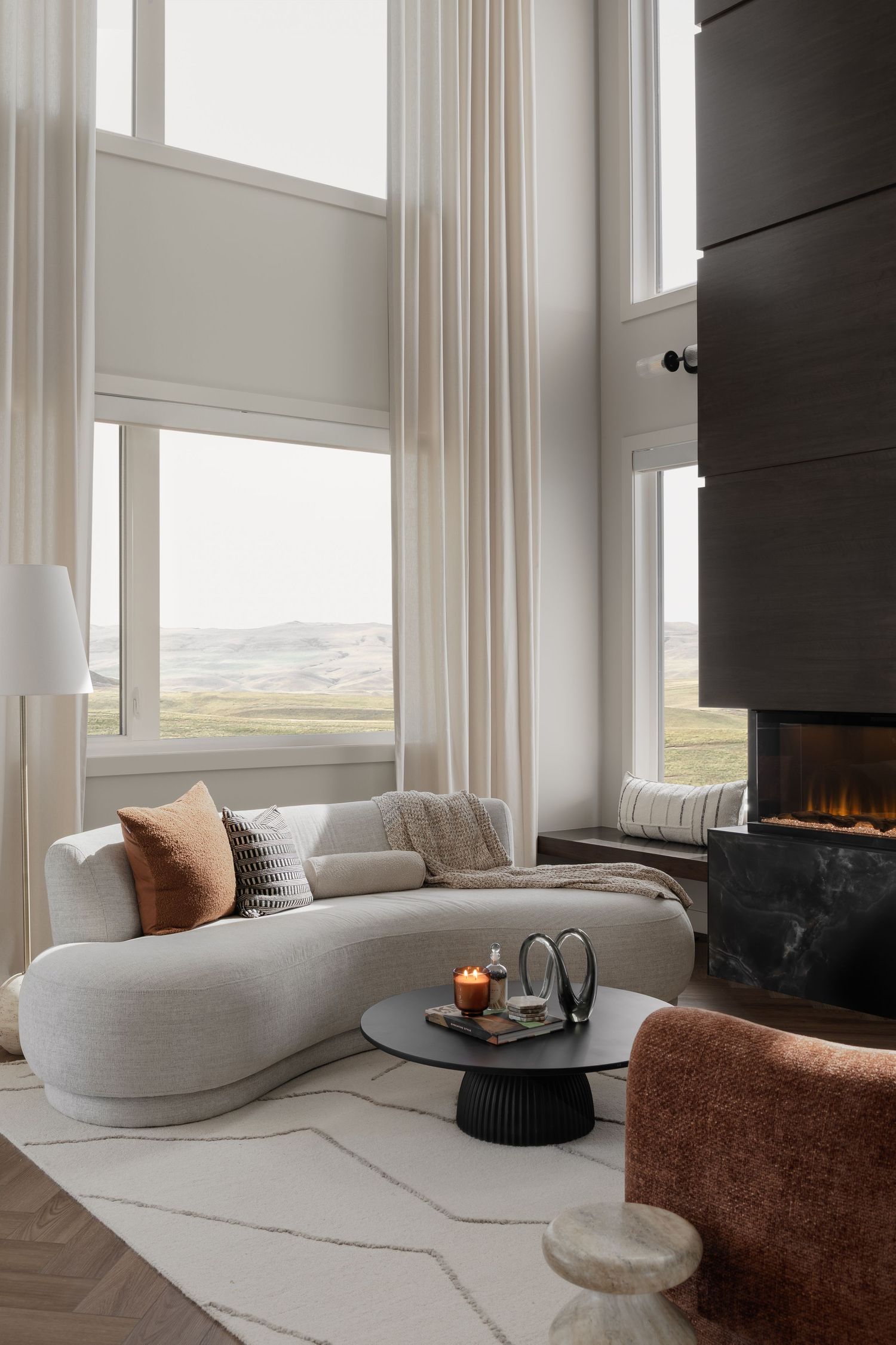 Luxurious living room with curved gray sofa, double-height windows, modern fireplace, and scenic landscape views.