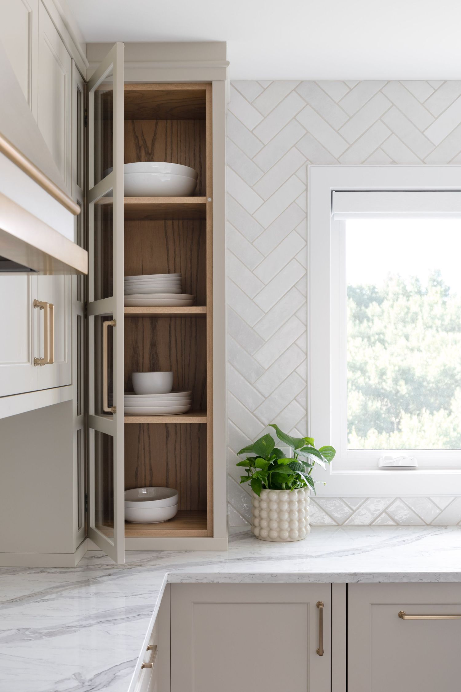 Open kitchen cabinet with warm wood interior displaying stacked white dishes beside herringbone tile backsplash and marble countertop.