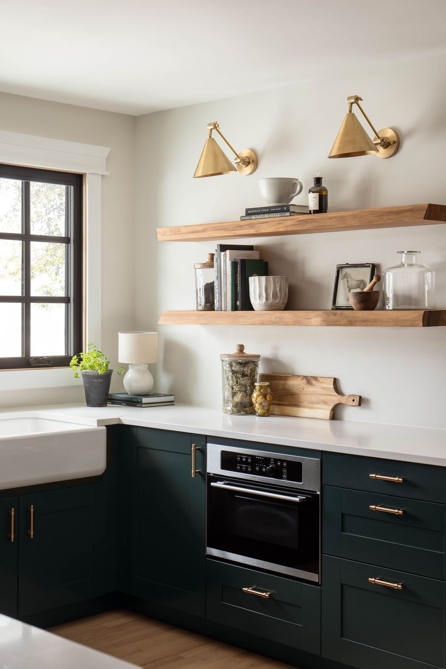 Dark green shaker kitchen cabinets with brass hardware, white quartz counters, farmhouse sink, and open wood shelves.
