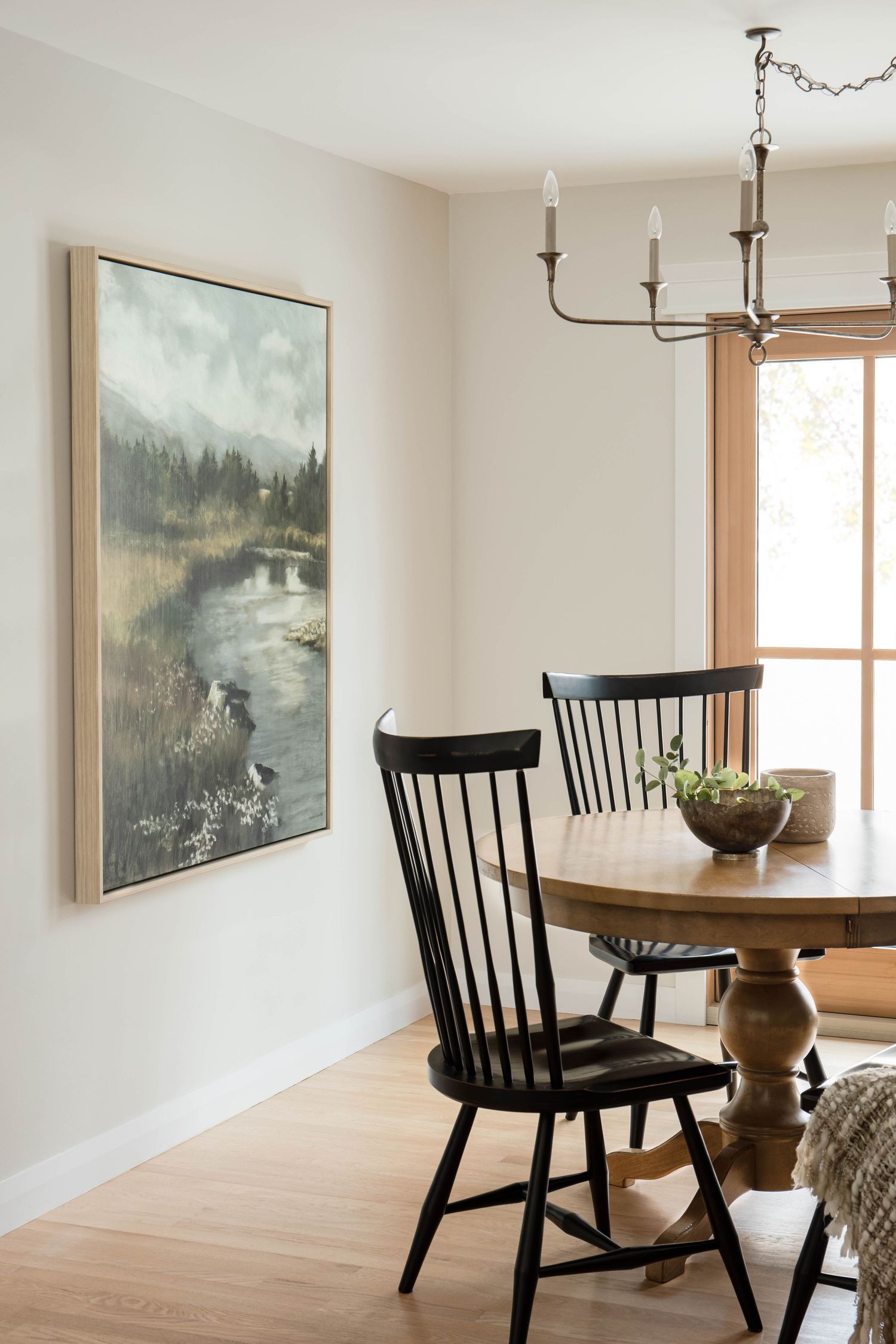 Dining nook with round wood table, black Windsor chairs, brass chandelier, and large landscape painting on wall.