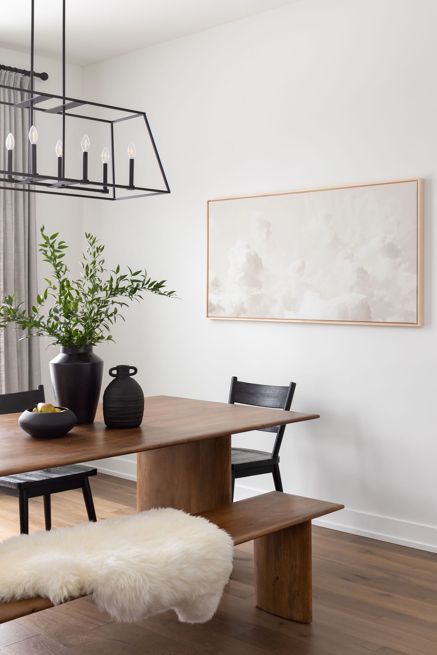 Modern dining room with walnut table, black chairs, sheepskin bench, dark vases, and abstract art on white wall.