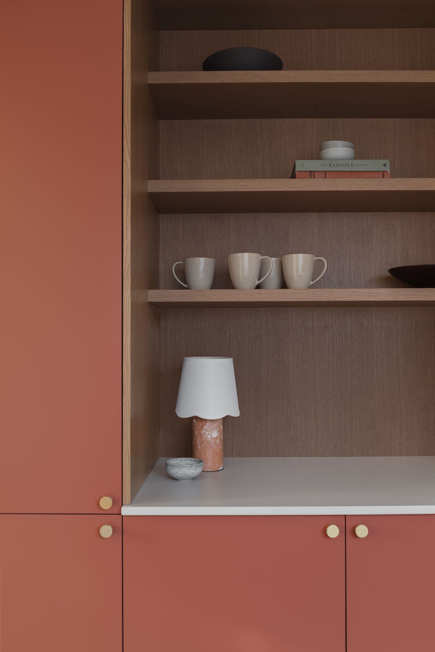 Terracotta pink cabinetry with open oak shelves displaying ceramic mugs, bowls, and a small table lamp.