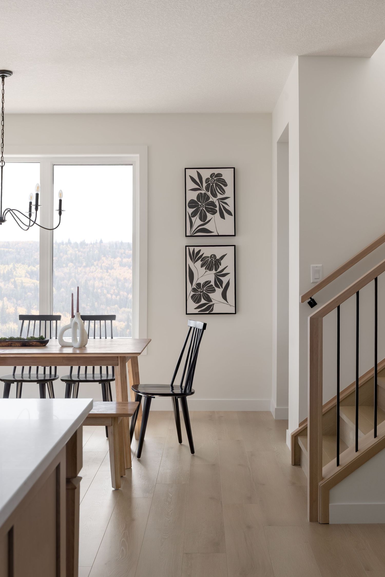 Modern dining room with black Windsor chairs, botanical wall art, staircase, and forest view window.