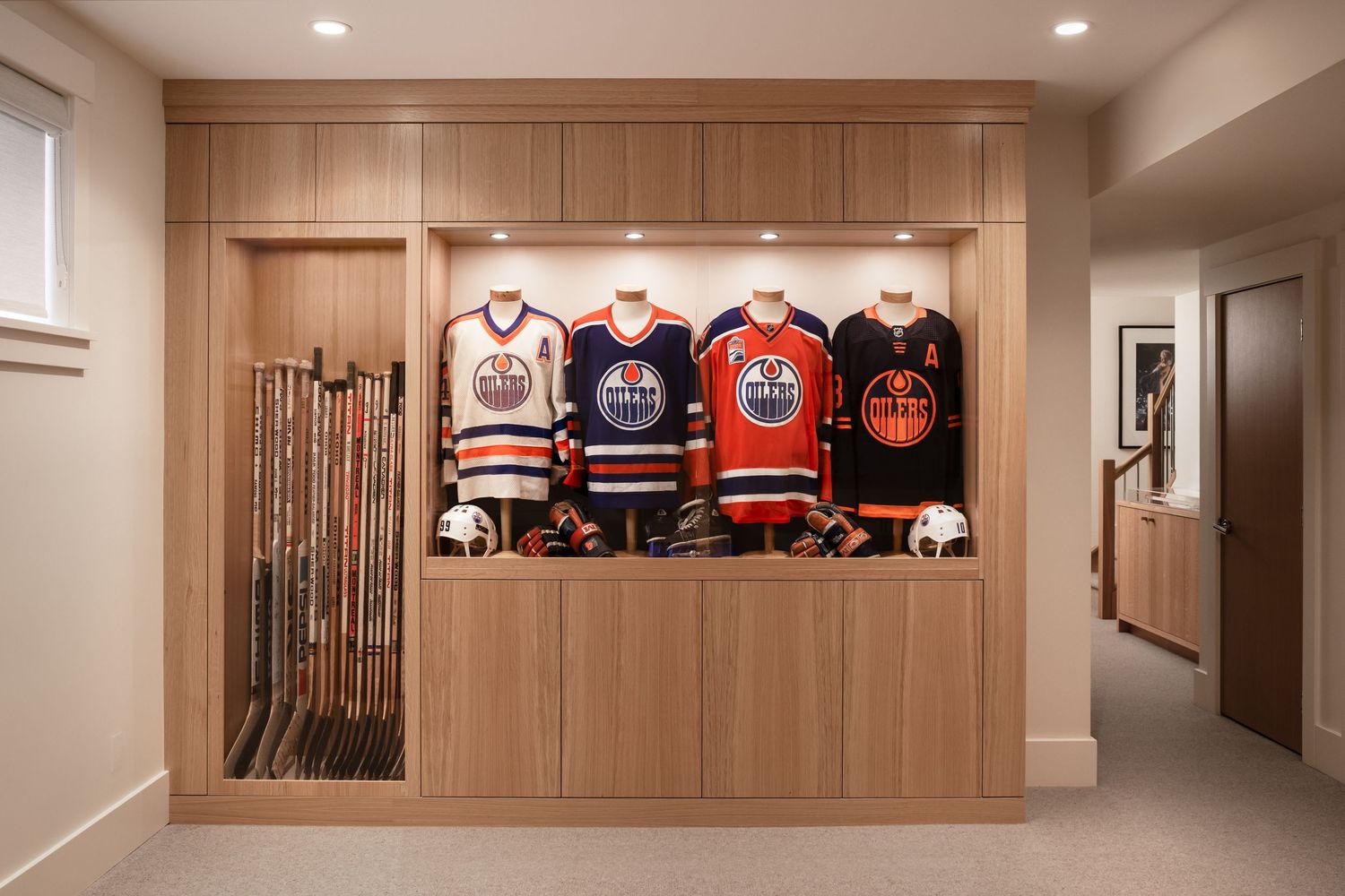 Custom wood built-in display case showcasing Edmonton Oilers hockey jerseys and sticks in a home hallway.