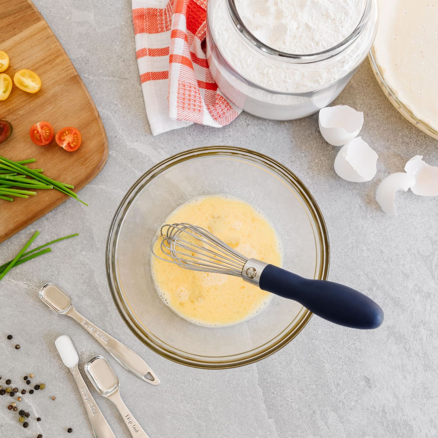 Glass mixing bowl with whisked eggs and a blue-handled whisk surrounded by baking ingredients.