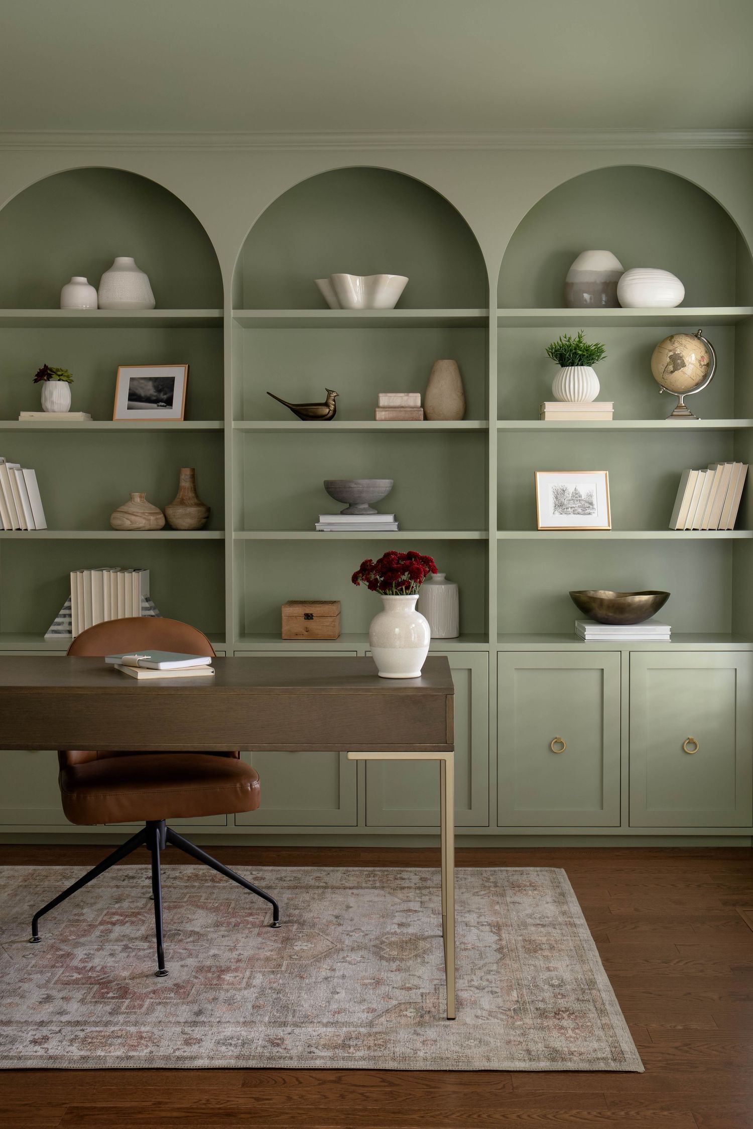 Sage green home office with arched built-in shelving, wood desk, leather chair, and curated decor accessories.