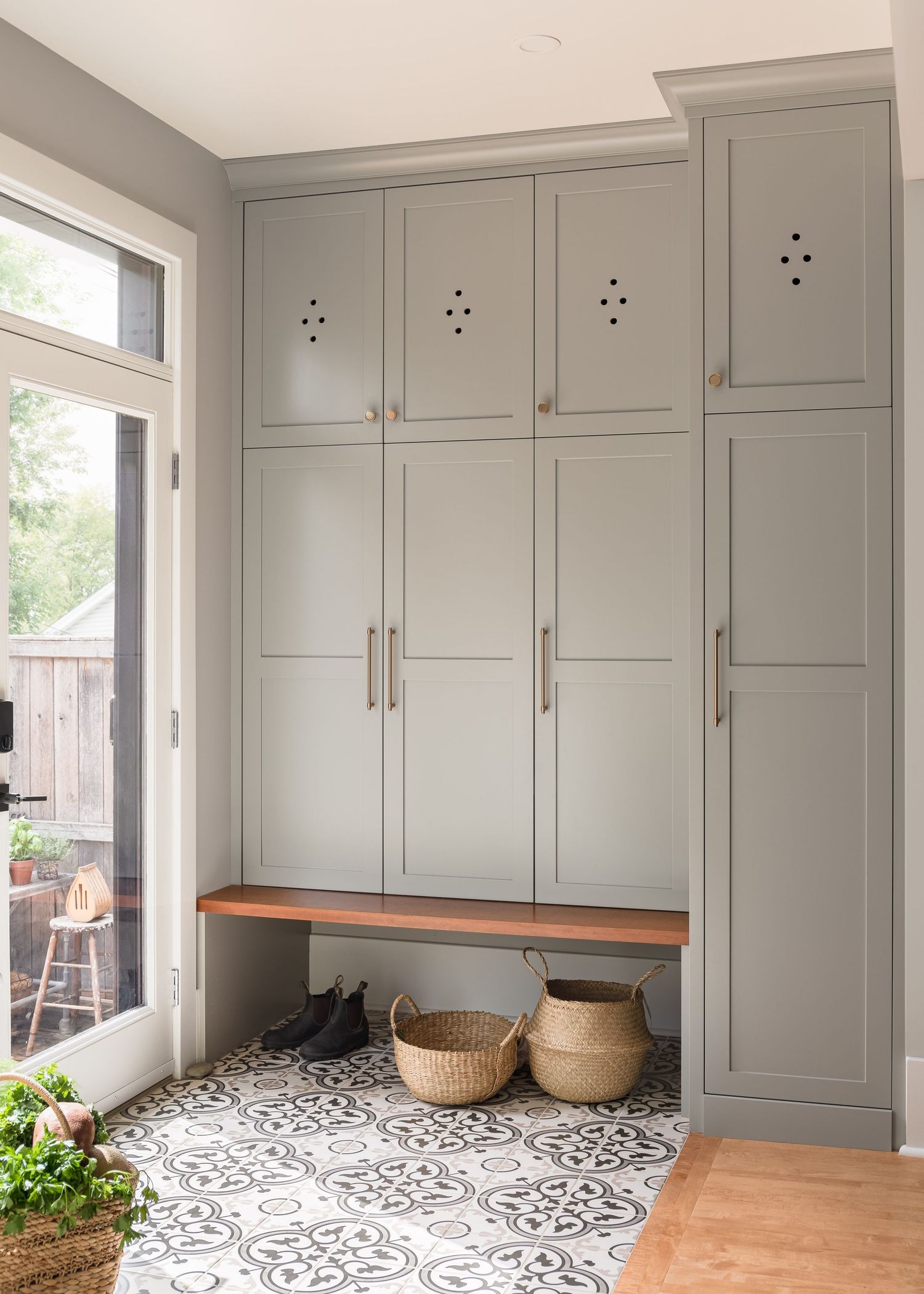 Gray shaker-style mudroom built-in lockers with wood bench, patterned tile floor, woven baskets, and glass door to outdoor patio.