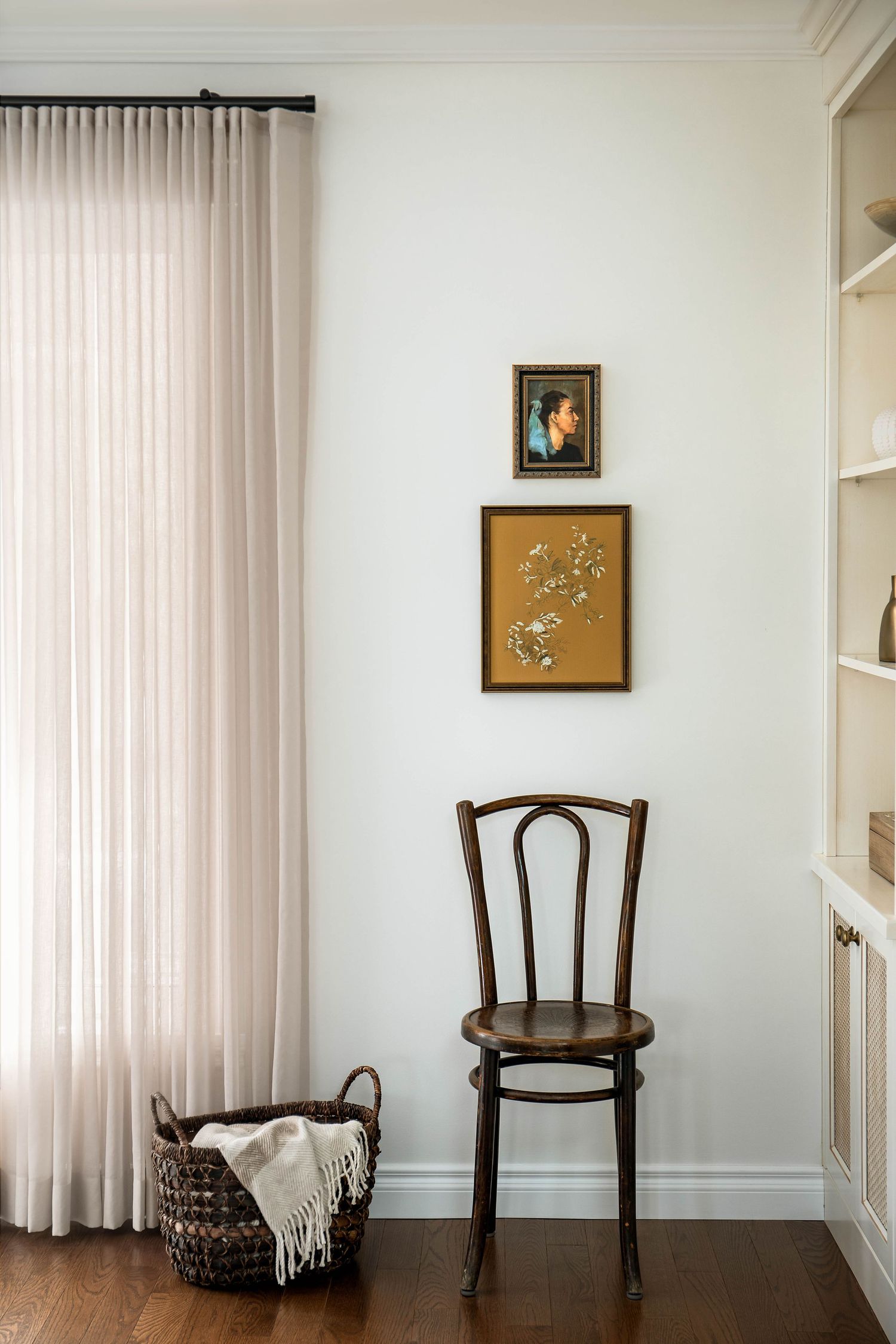 Minimalist corner with bentwood chair, woven basket, sheer curtains, and two framed artworks on white wall.
