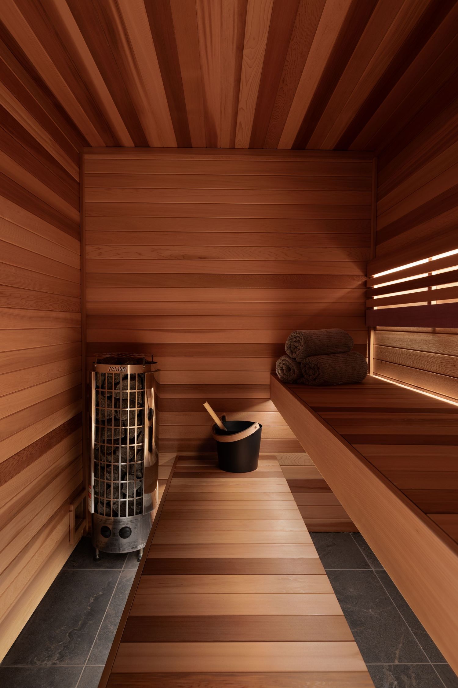 Cedar wood sauna with stone floor, cylinder heater, and built-in benches with rolled towels by Calgary Interior photographer Rebecca Frick.