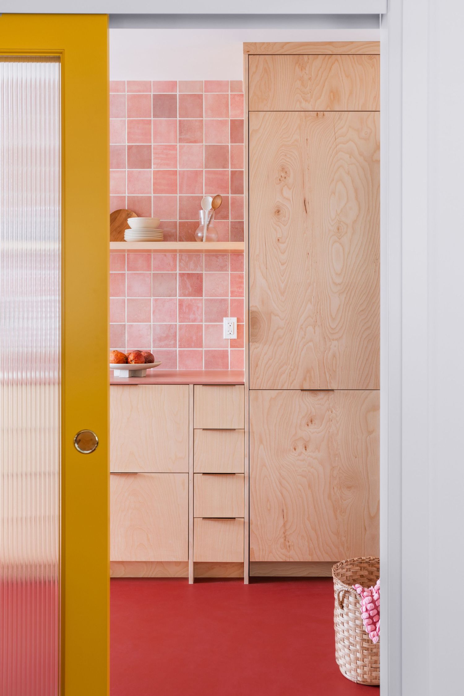 Colorful kitchen with pink square tile backsplash, light plywood cabinets, red floor, and yellow door frame.