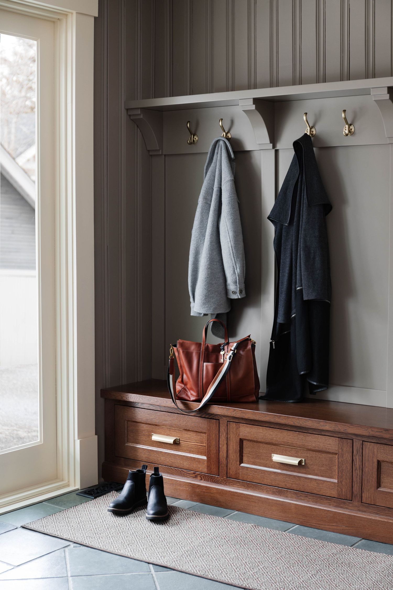 Mudroom entryway with dark beadboard walls, walnut storage bench, brass hooks, and hanging coats.