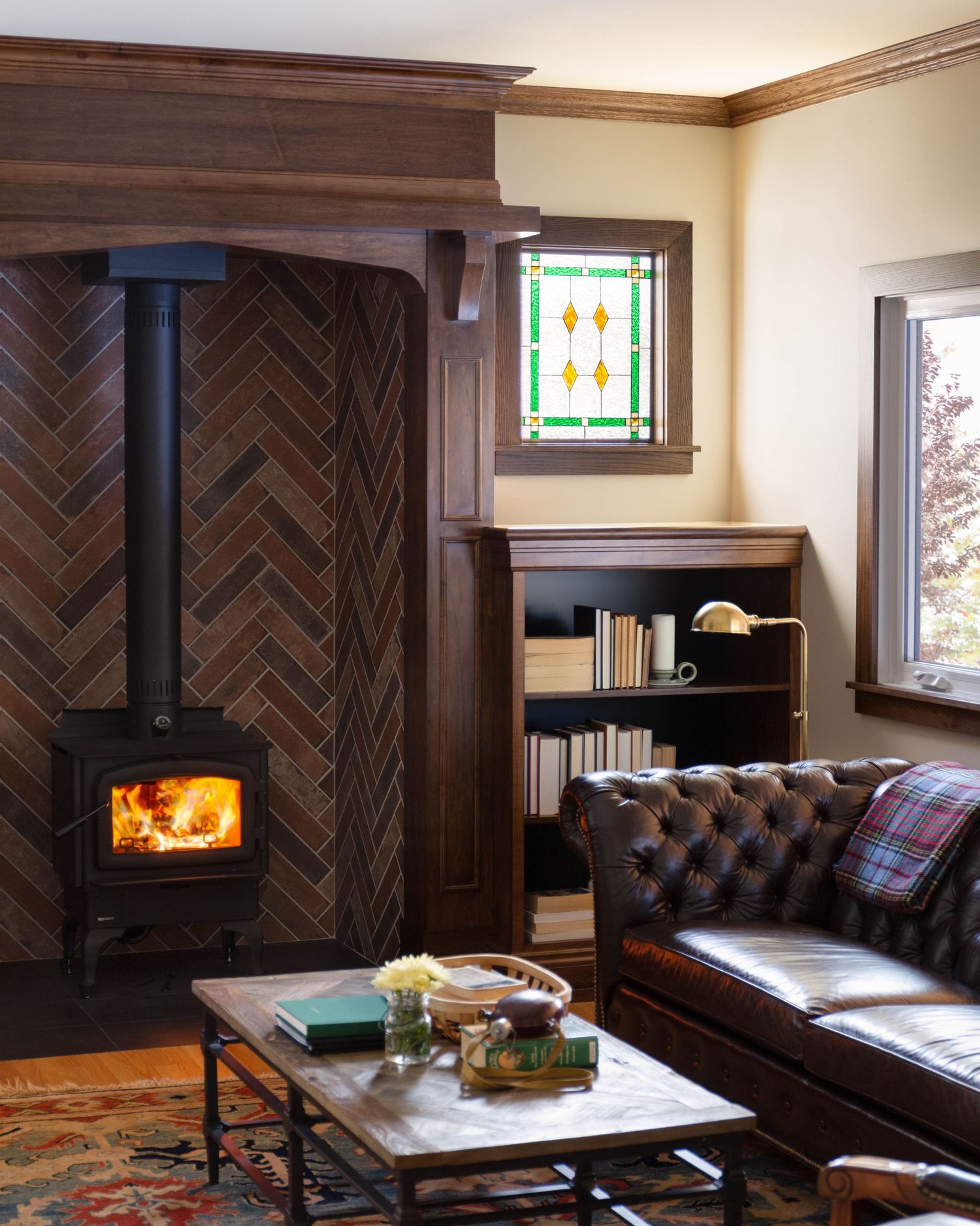 Cozy living room with wood-burning stove, herringbone tile wall, tufted leather sofa, stained glass window, and built-in bookcase.
