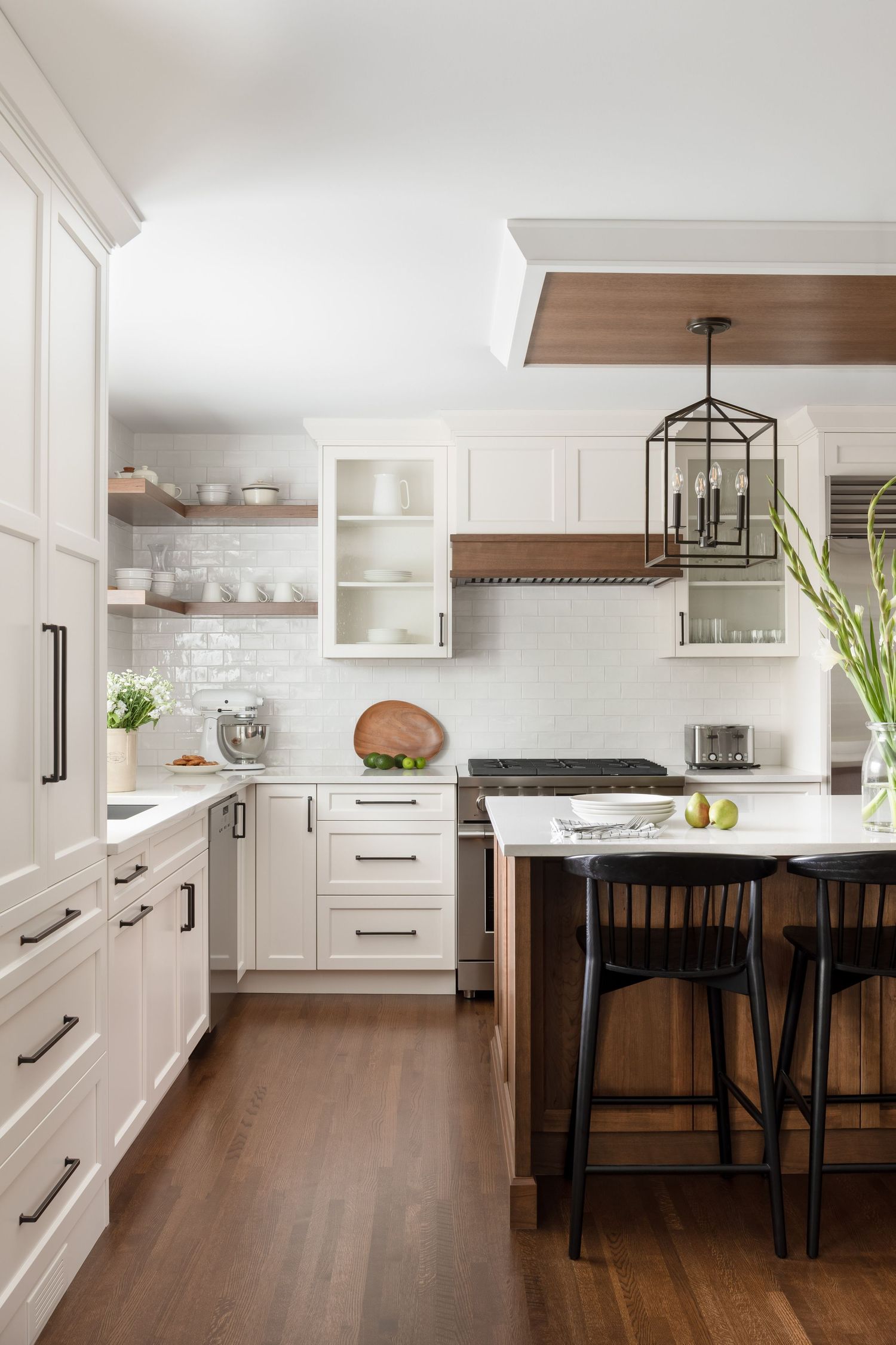 White kitchen with wood range hood, black island pendants, open shelving, and bar stools.